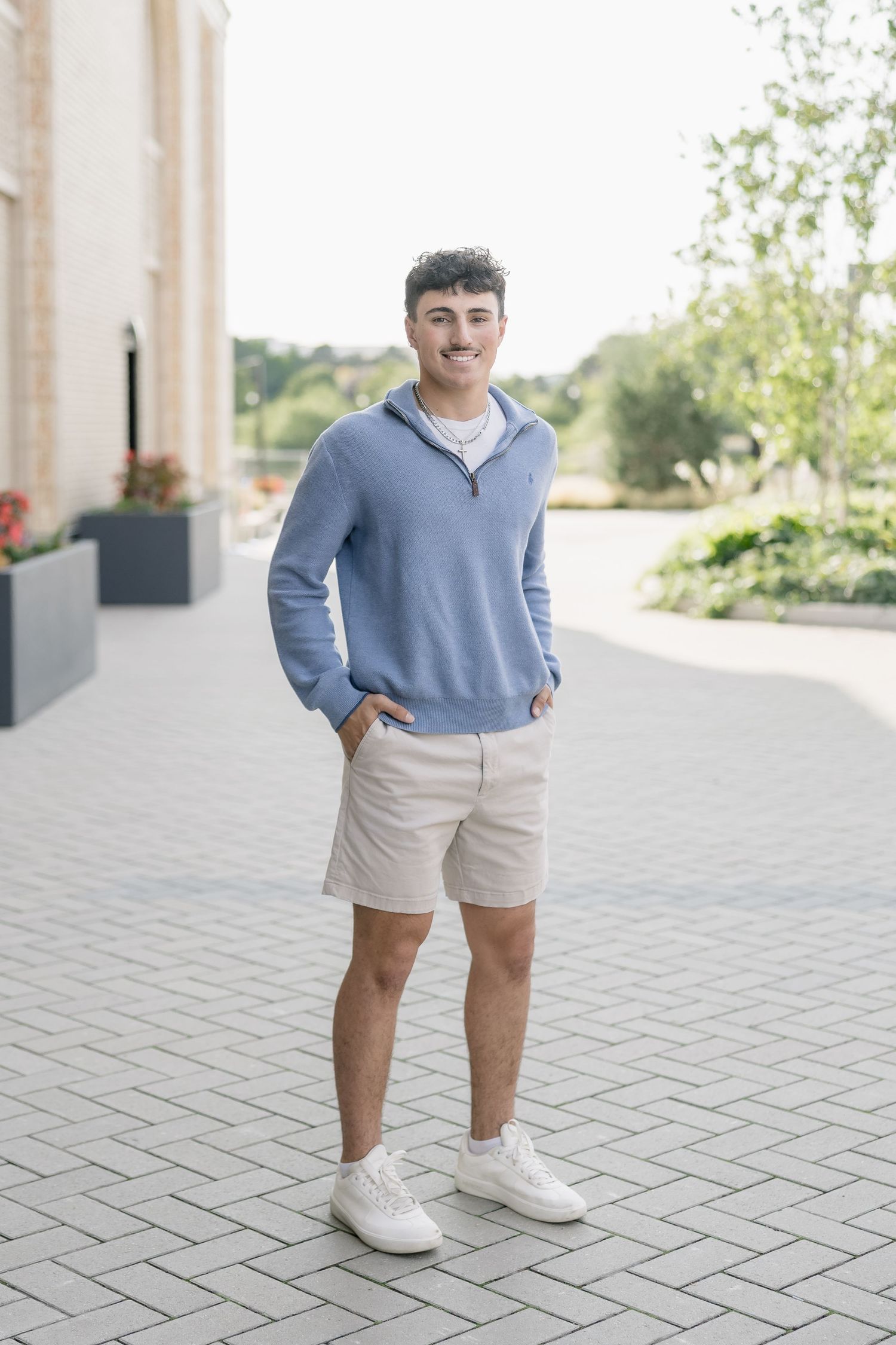 Person wearing a light blue sweater and khaki shorts stands on brick pavement with white sneakers.