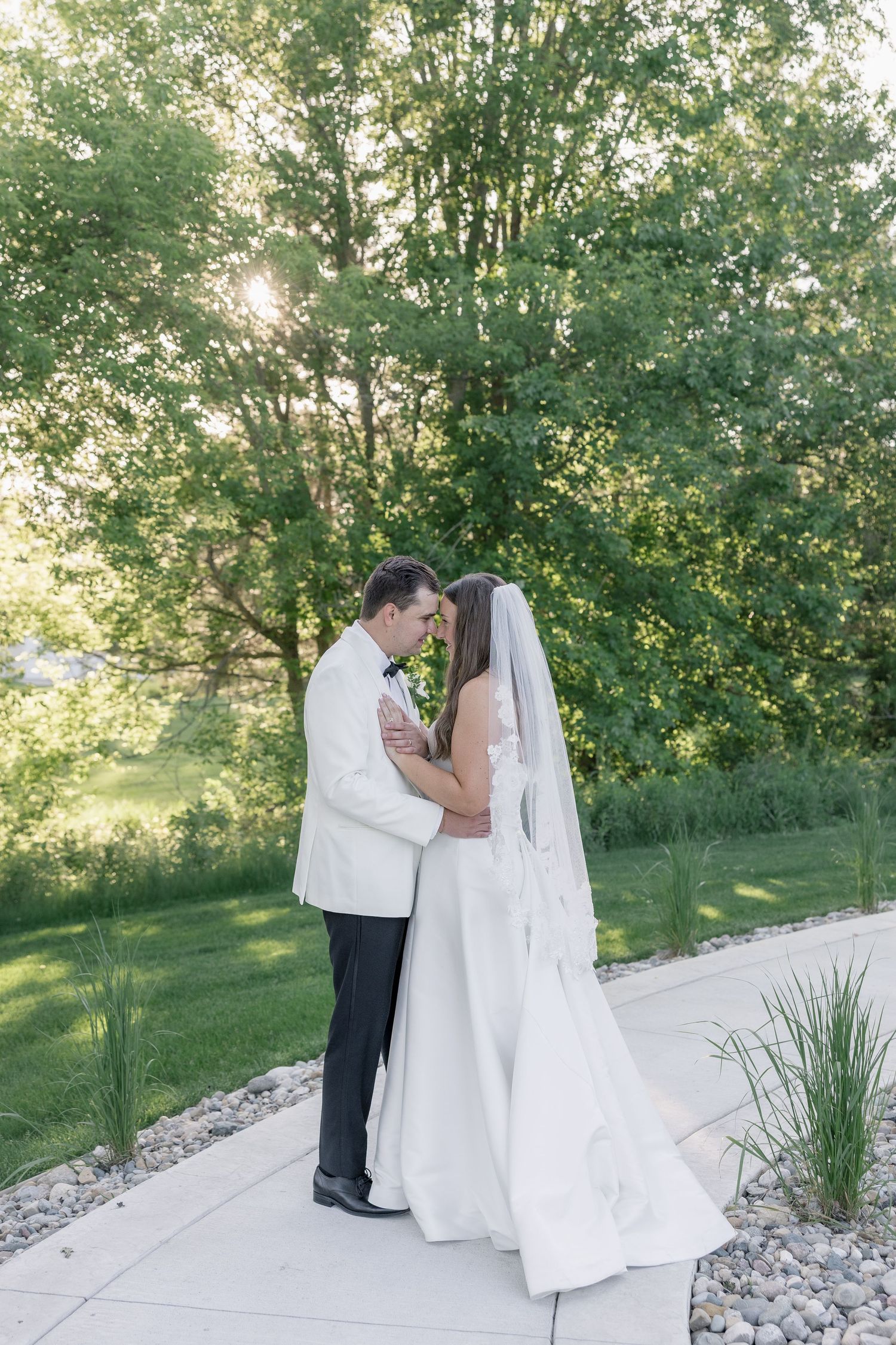 Bride and groom share a romantic forehead touch on a garden path, surrounded by lush green trees in soft sunlight.