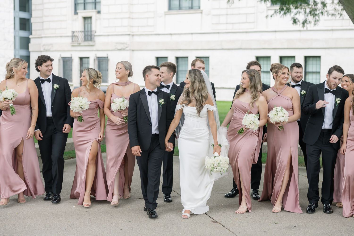 Wedding party in blush pink bridesmaid gowns and black tuxedos pose outdoors with bride and groom sharing a kiss.