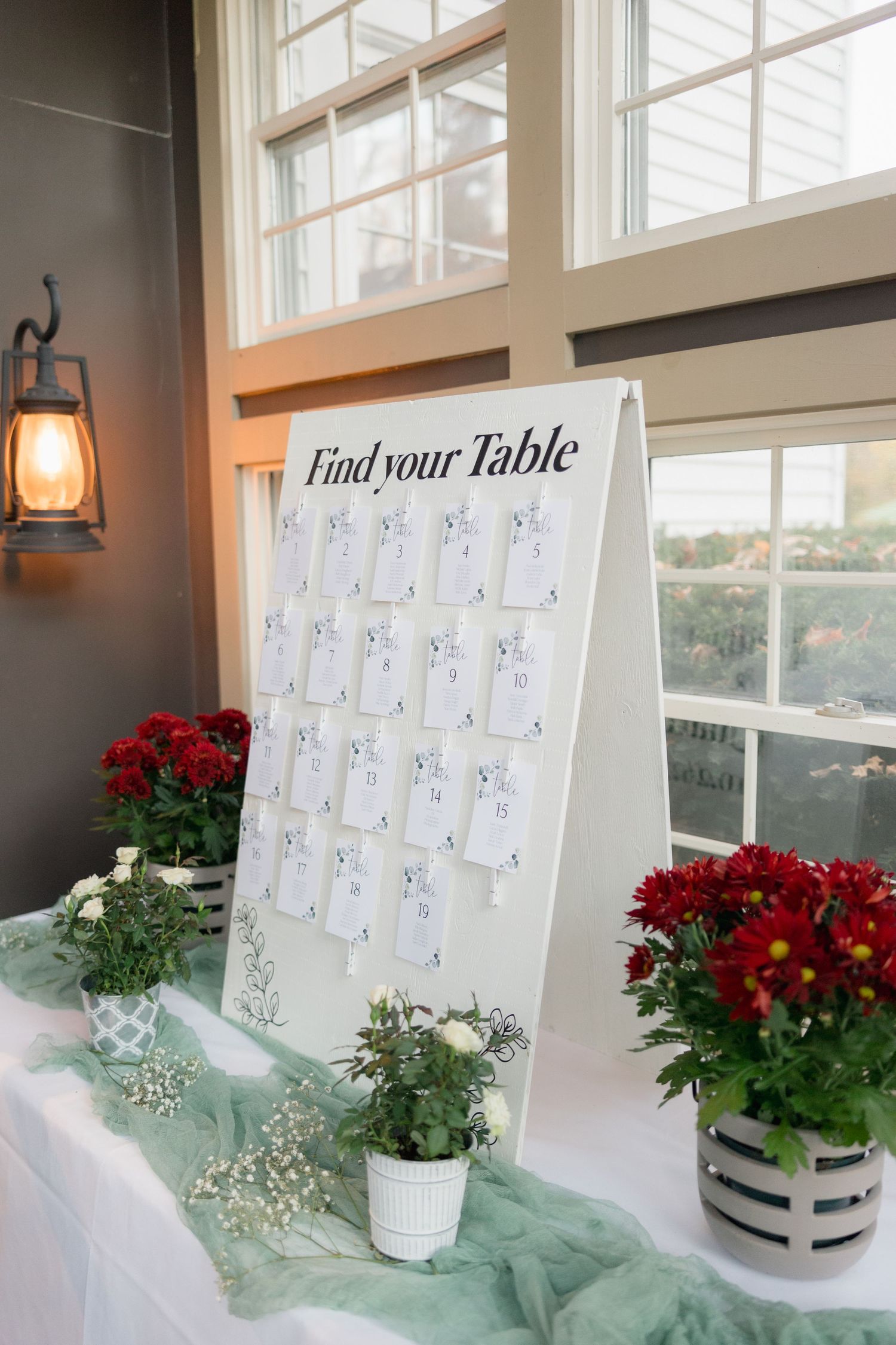 Wedding seating chart display with red flowers and greenery on mint-colored fabric.