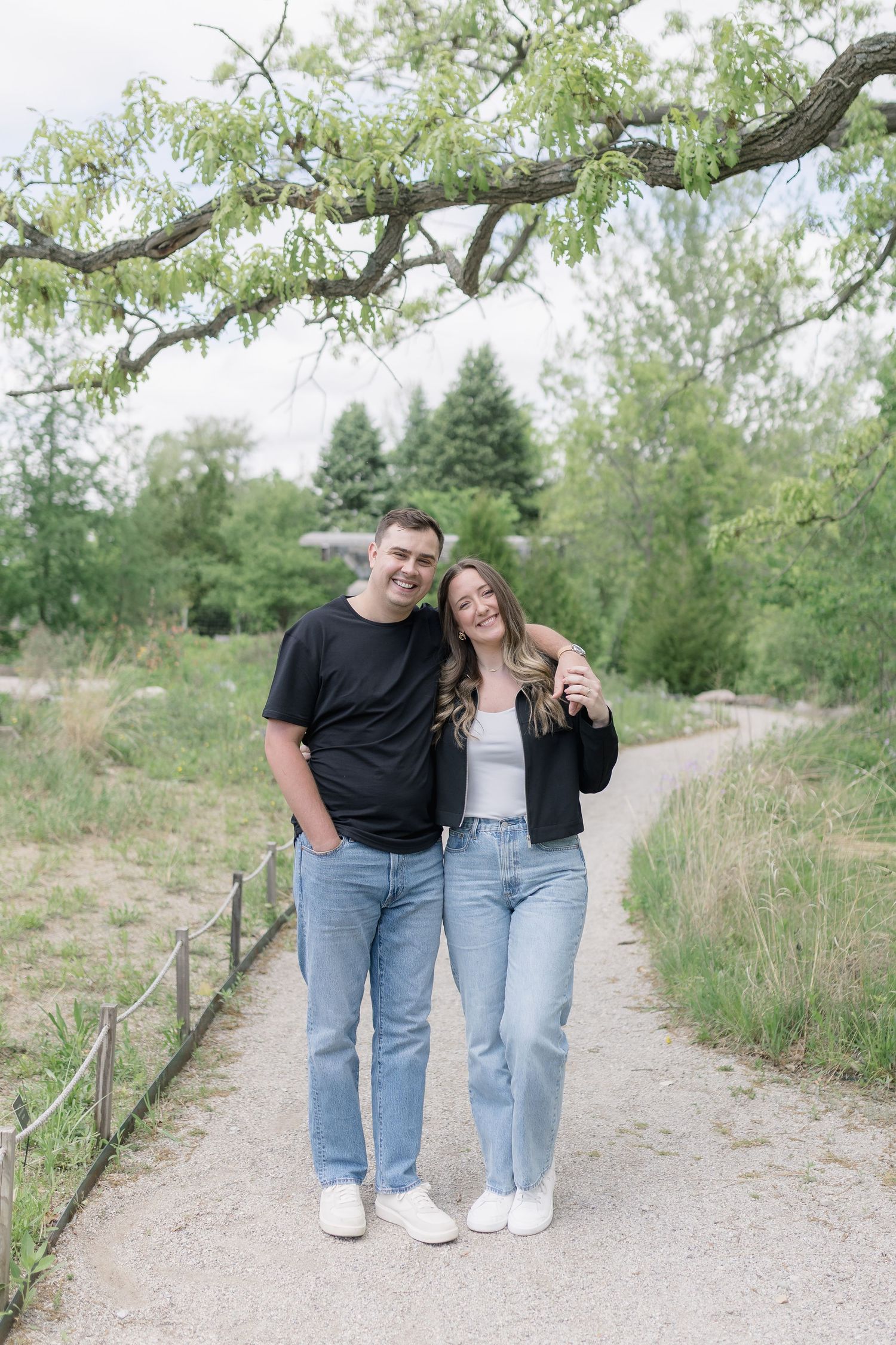 A couple walks together on a gravel path surrounded by green trees and natural scenery.