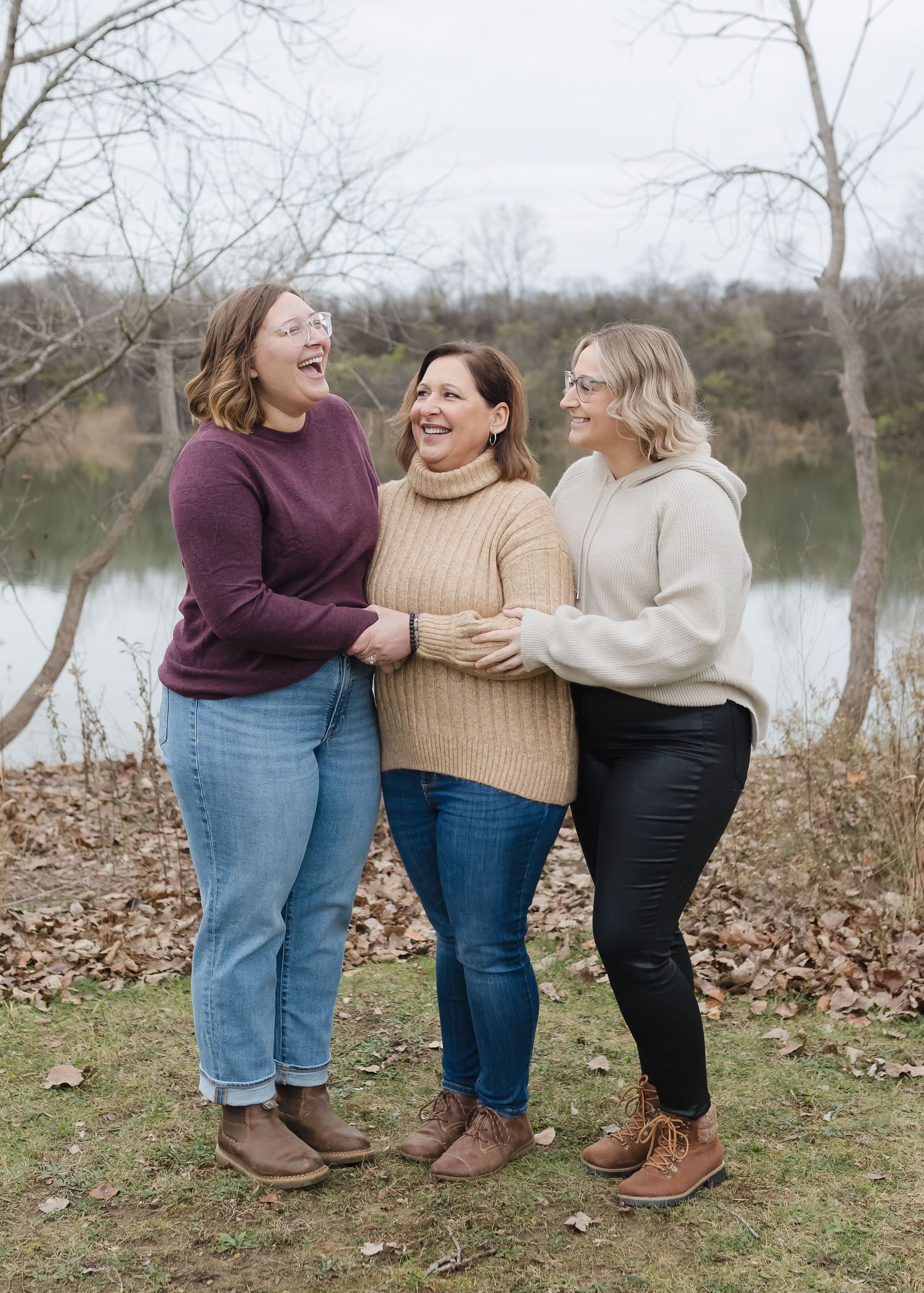 Three friends in casual fall outfits stand together by a lake, laughing and embracing on fallen autumn leaves.