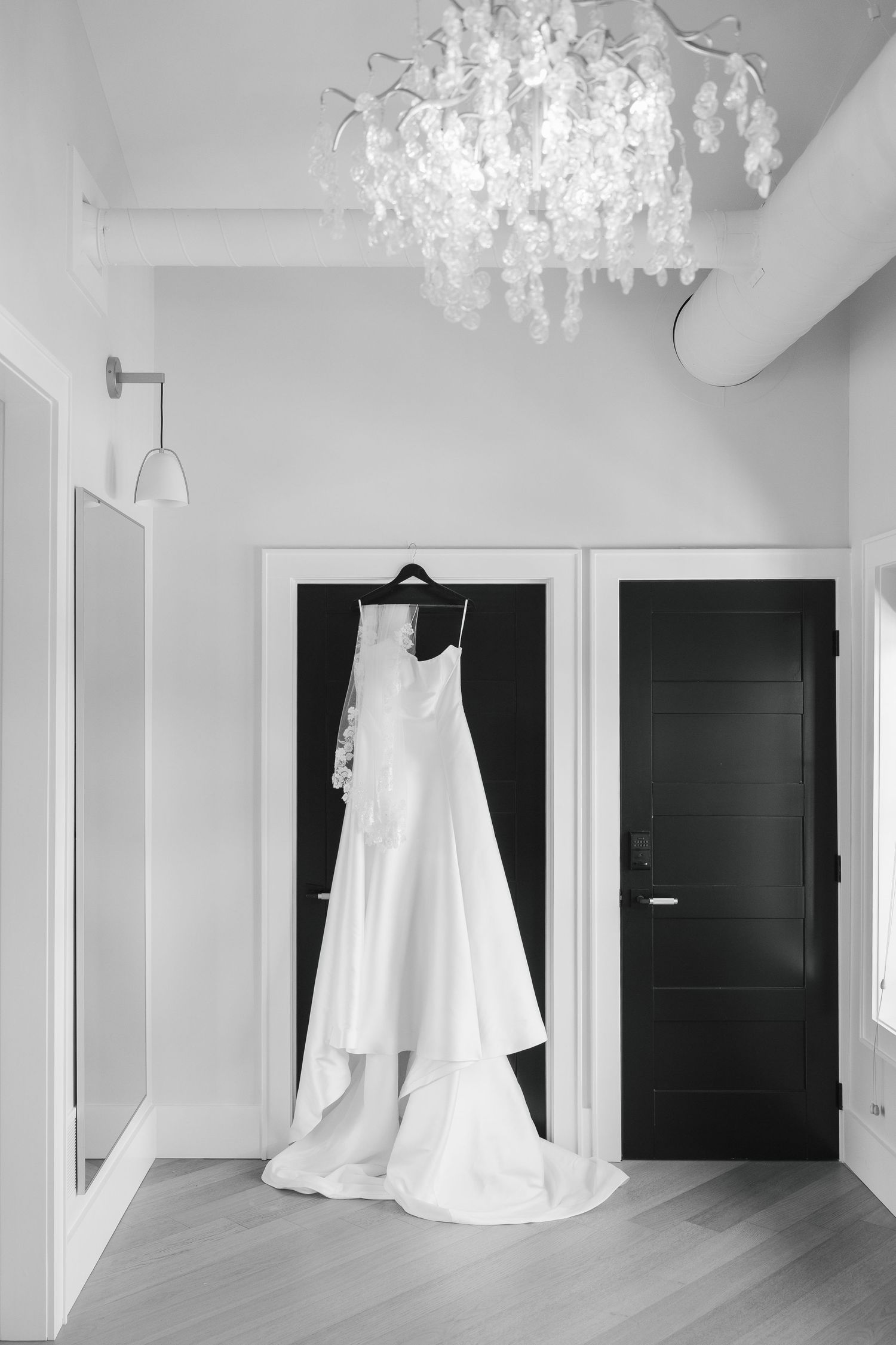 Elegant white wedding dress hanging on a hanger in a modern room with a floral chandelier, black and white photography.