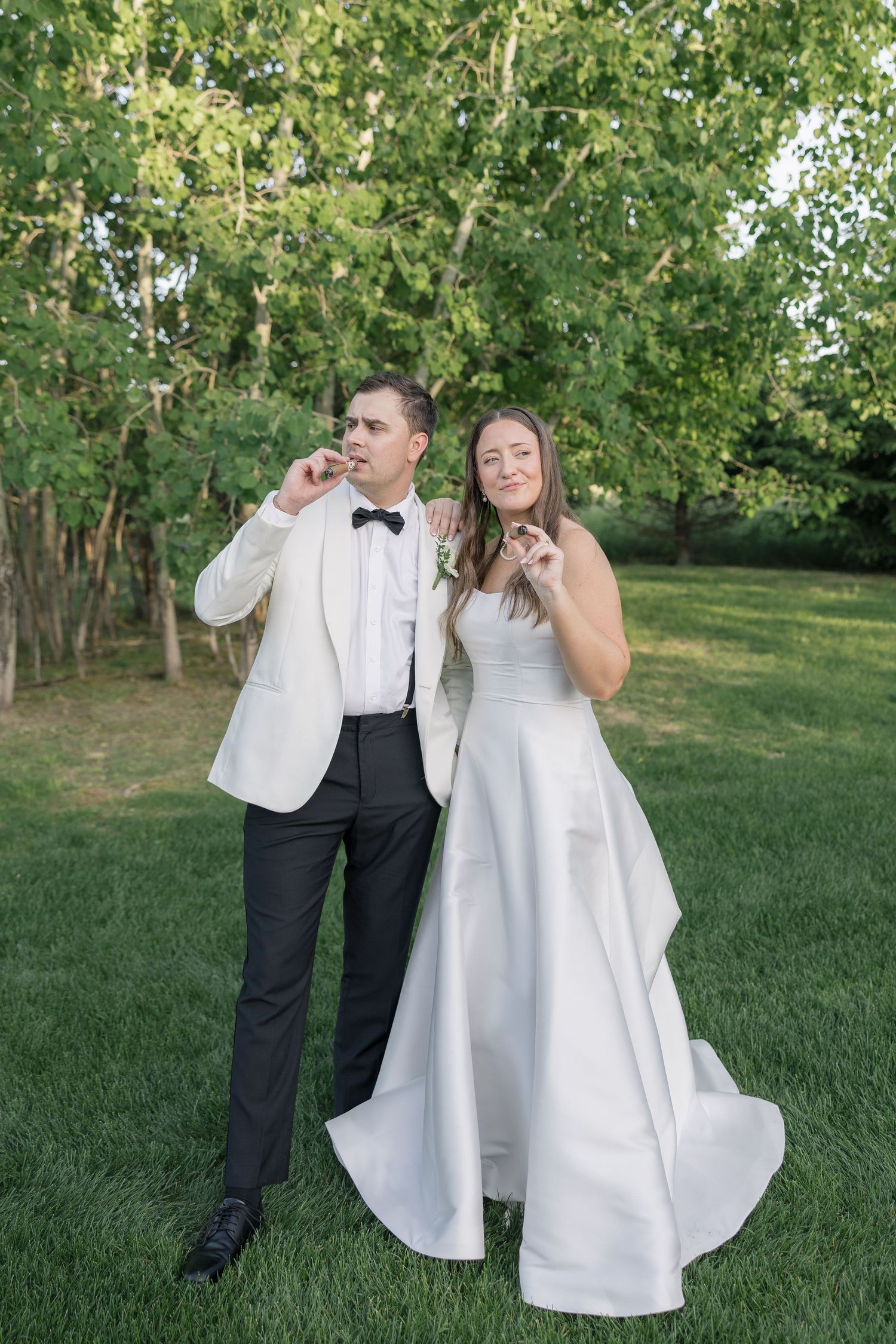 Bride and groom posing playfully outdoors on green grass, wearing wedding attire, surrounded by lush trees.
