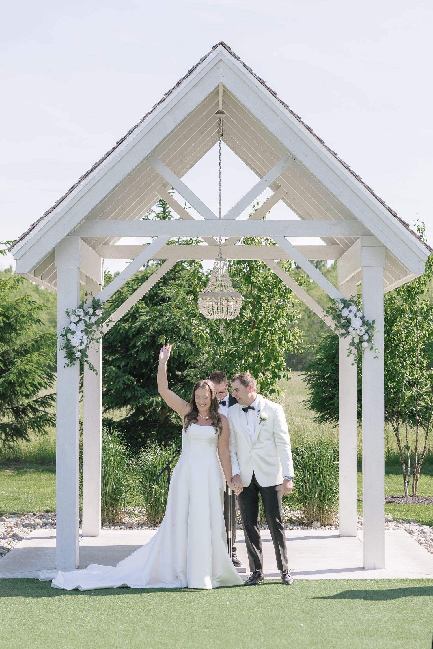Bride and groom standing under a white wooden gazebo with floral accents and chandelier on a sunny wedding day.