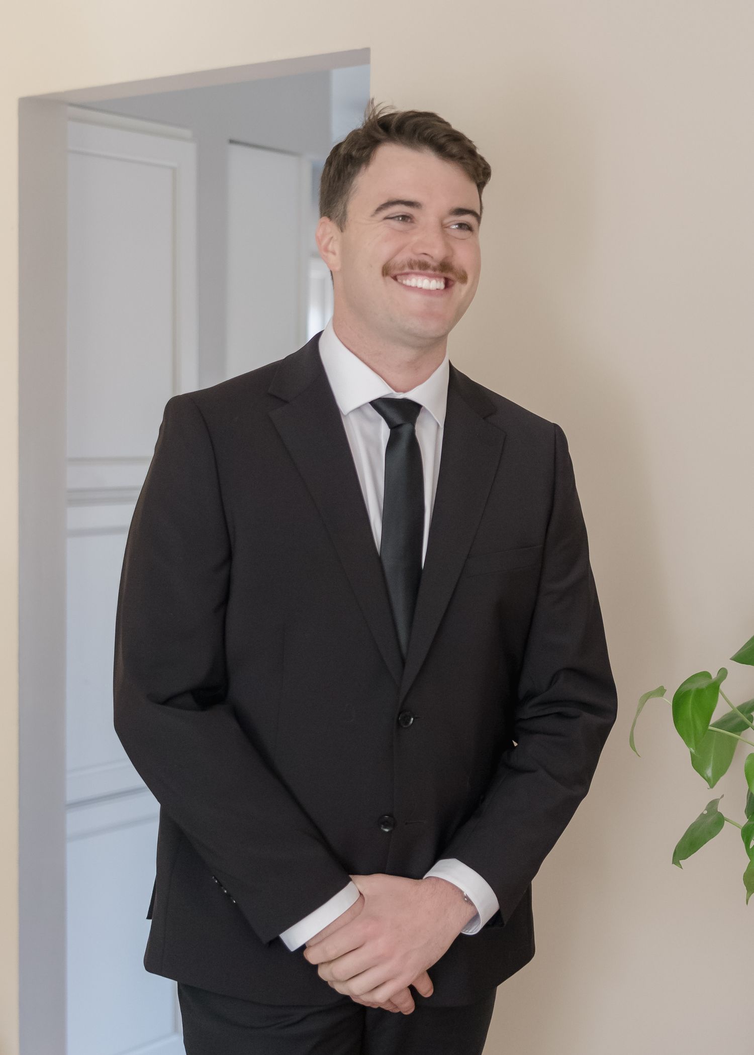 A portrait of a smiling groom in a classic black suit and tie against a light interior background.