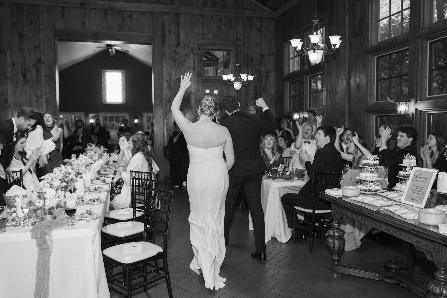 Black and white photo of wedding reception guests celebrating at elegant venue with warm lighting and table settings.