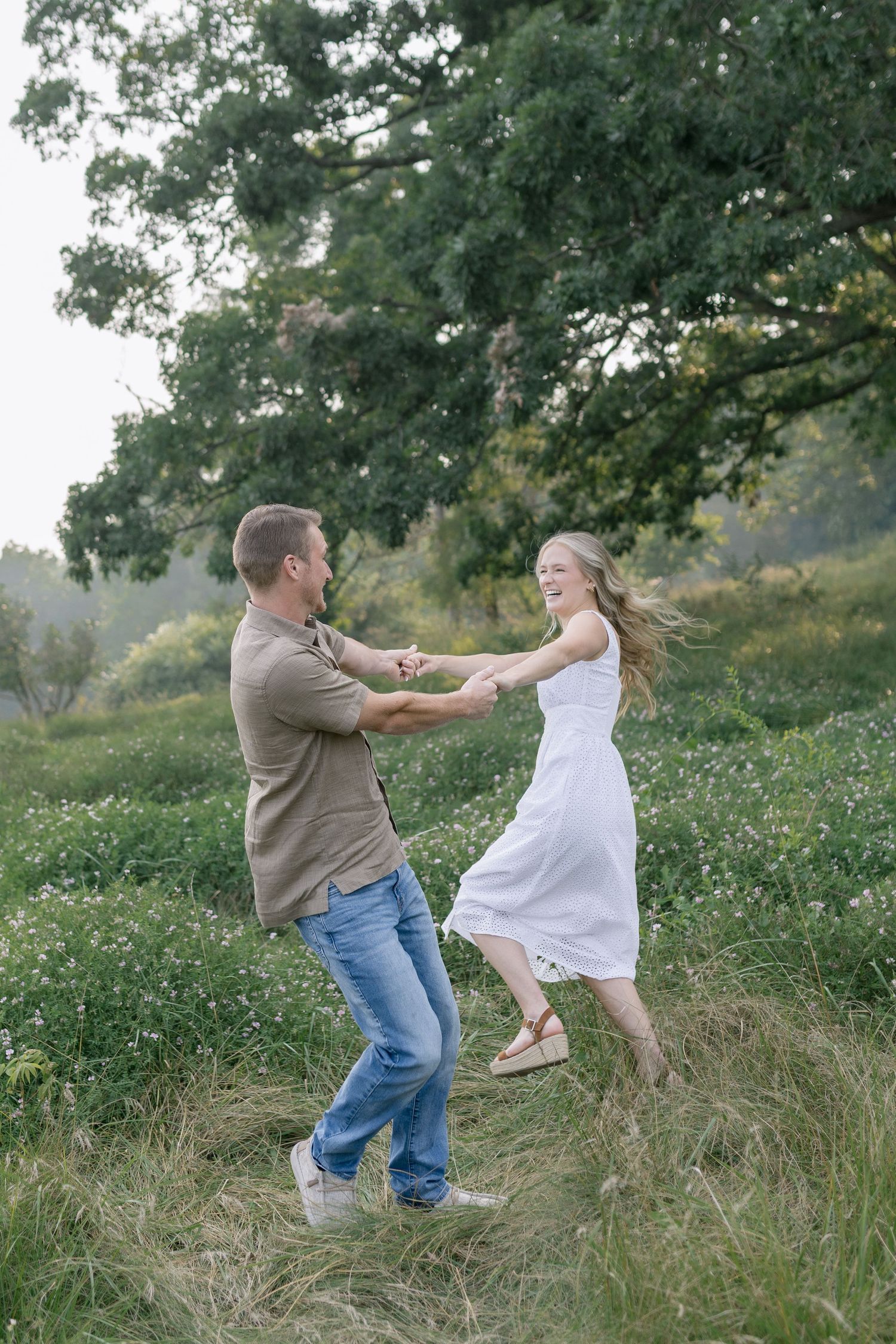 A couple dances playfully together in a meadow surrounded by trees on a summer afternoon.