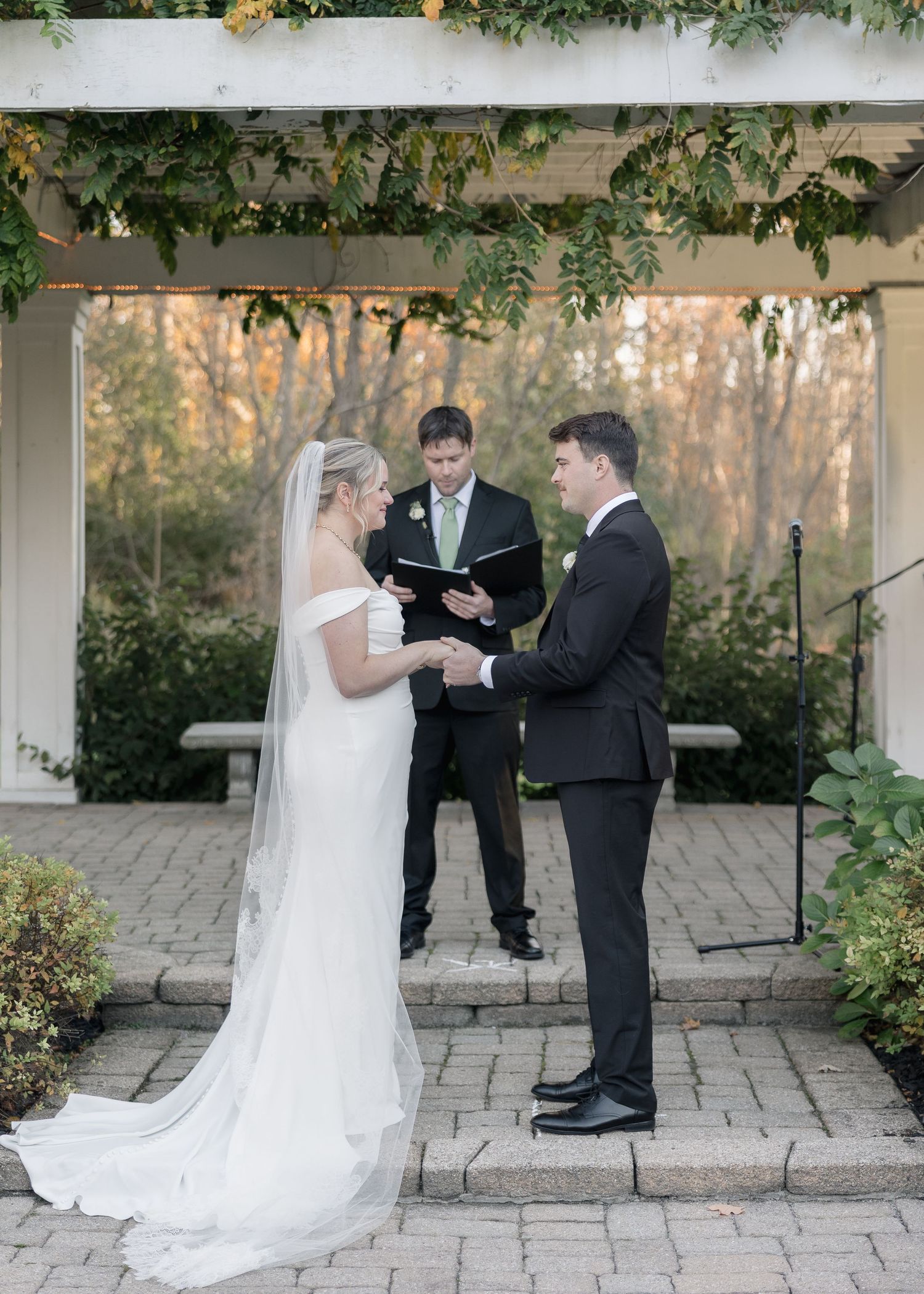 Wedding ceremony takes place beneath a white pergola decorated with greenery during autumn.