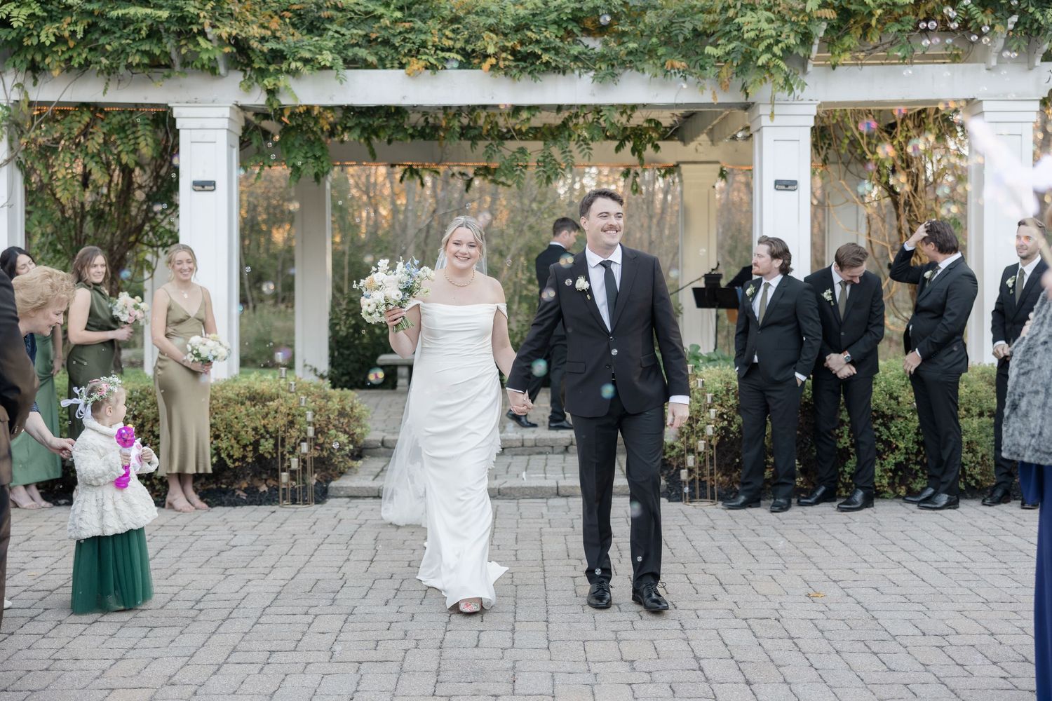 A wedding celebration with guests gathered outdoors under a decorated pergola.