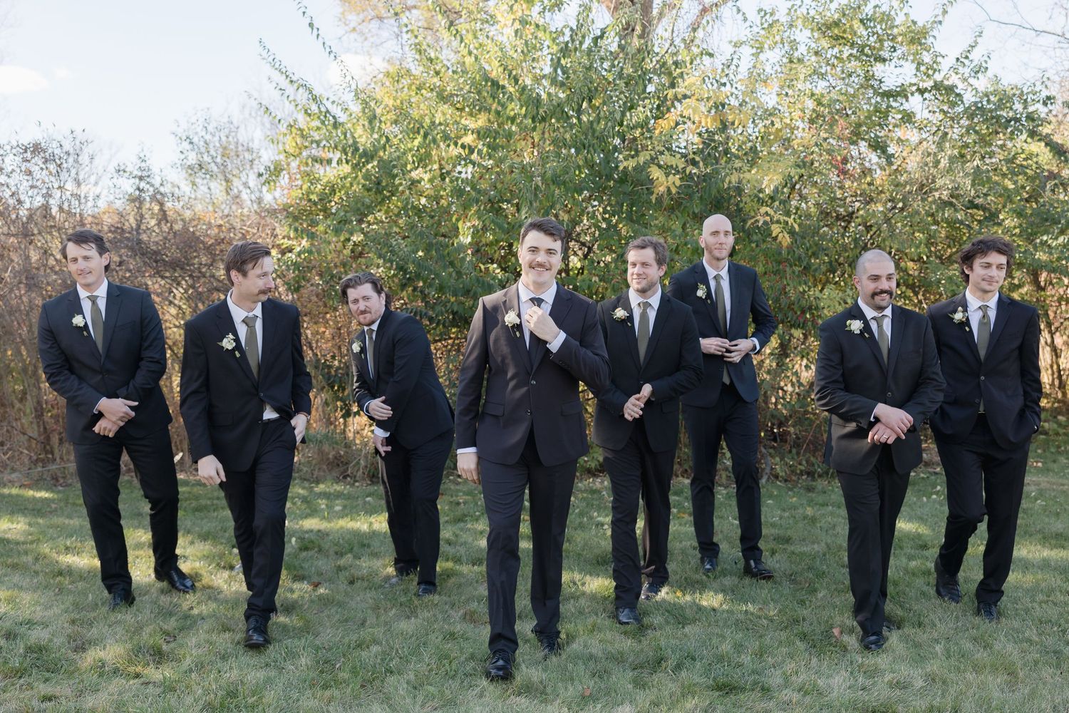 A formal wedding party of groomsmen in black suits stand outdoors in front of autumn foliage.