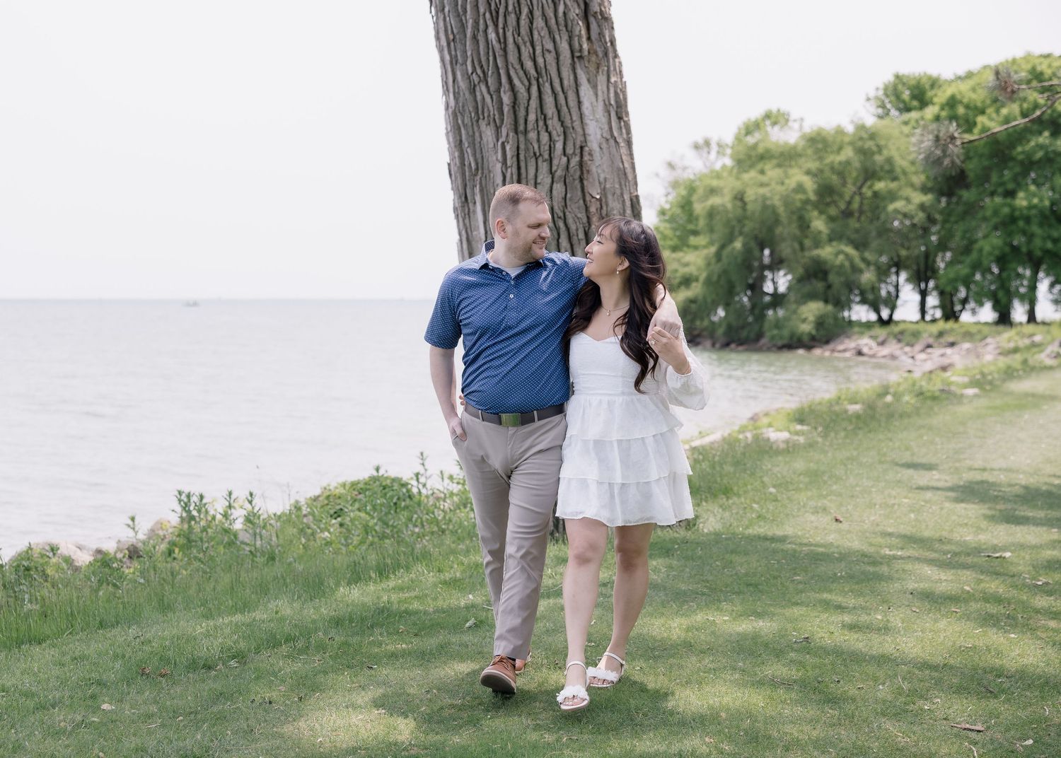 A romantic couple embraces while standing in a green park setting with trees in the background.