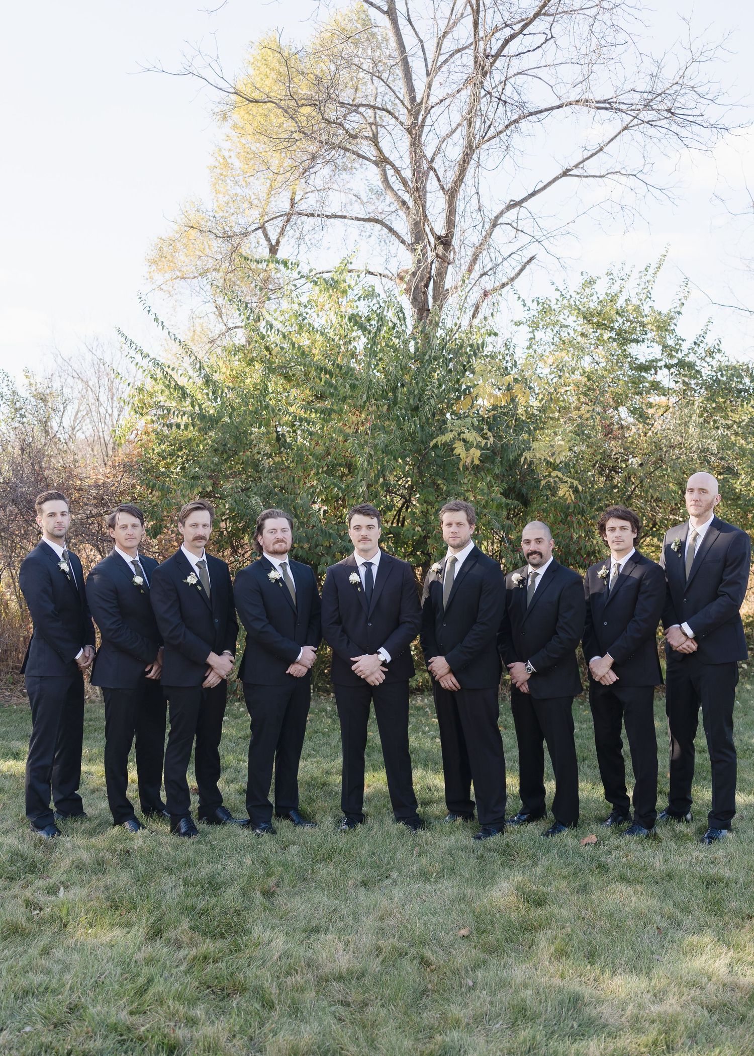 A wedding party in black suits stands in a line outdoors against an autumn backdrop with trees and grass.