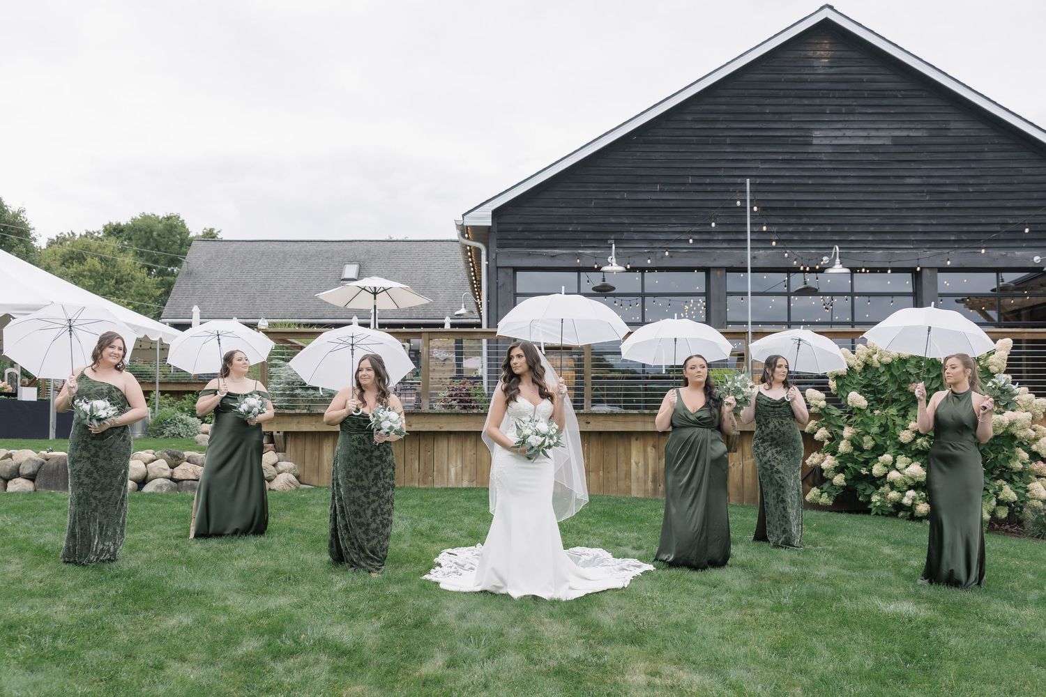 Wedding party holding white umbrellas against dark barn in garden setting.