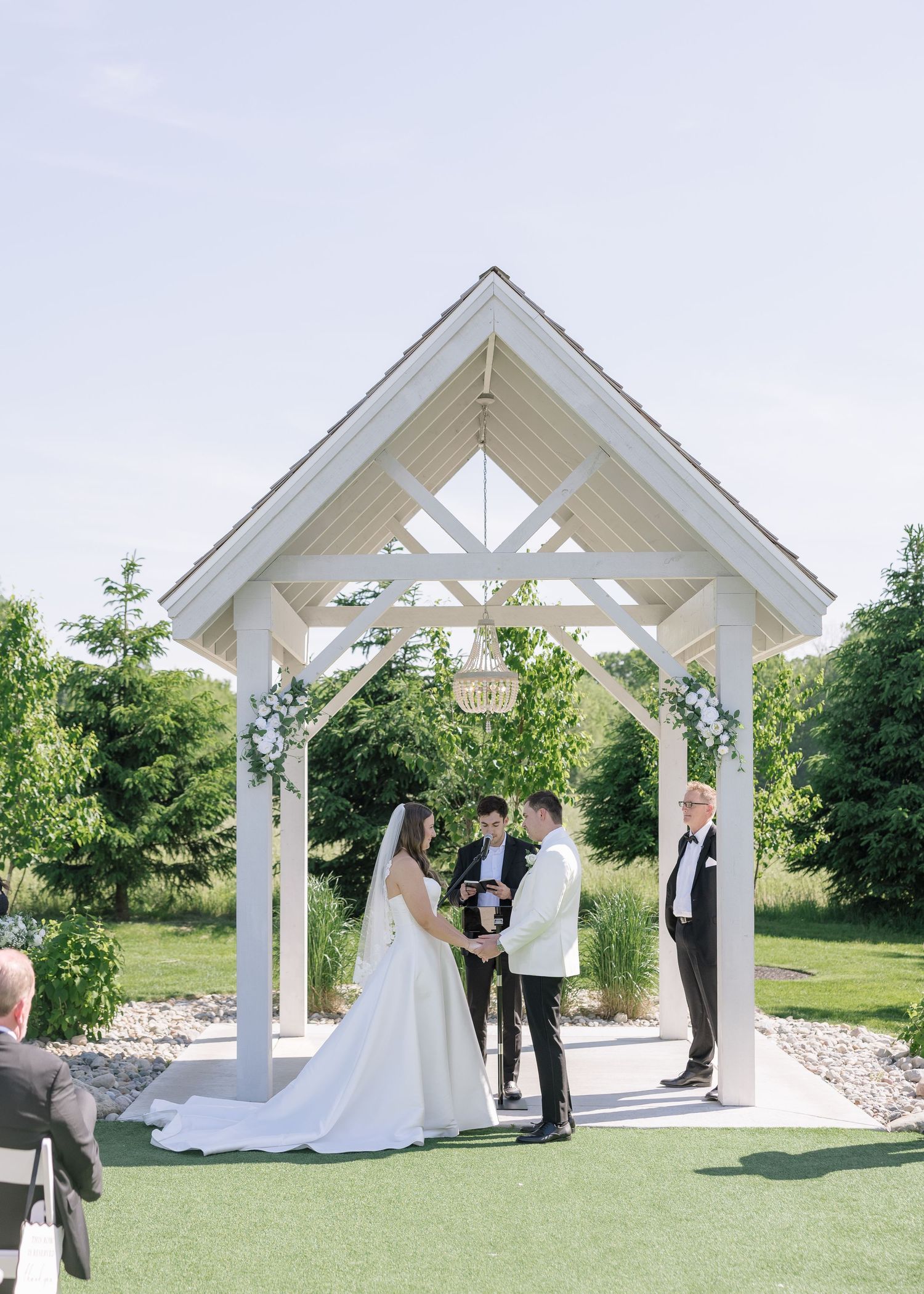 Couple exchanges vows under white garden gazebo decorated with flowers at an outdoor wedding ceremony.