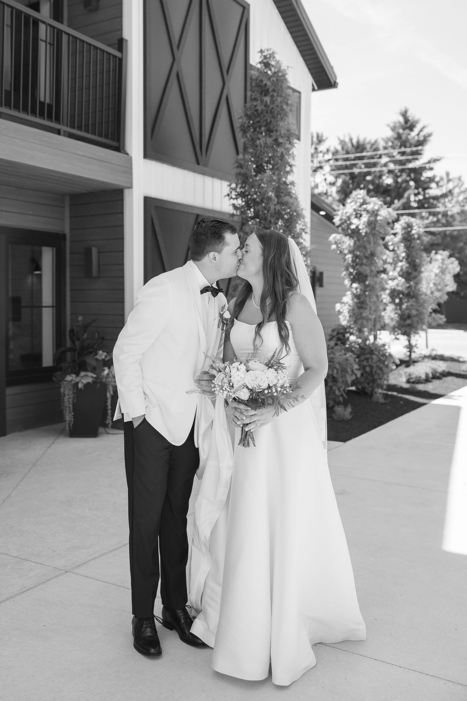 Black and white photo of a bride and groom sharing a kiss outside a modern barn venue on their wedding day.