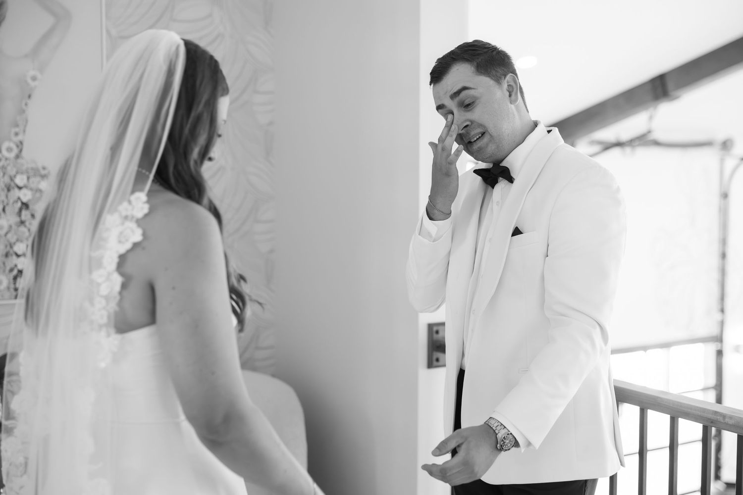 Emotional groom wiping tears during wedding first look with bride in white gown and veil, black and white photo.