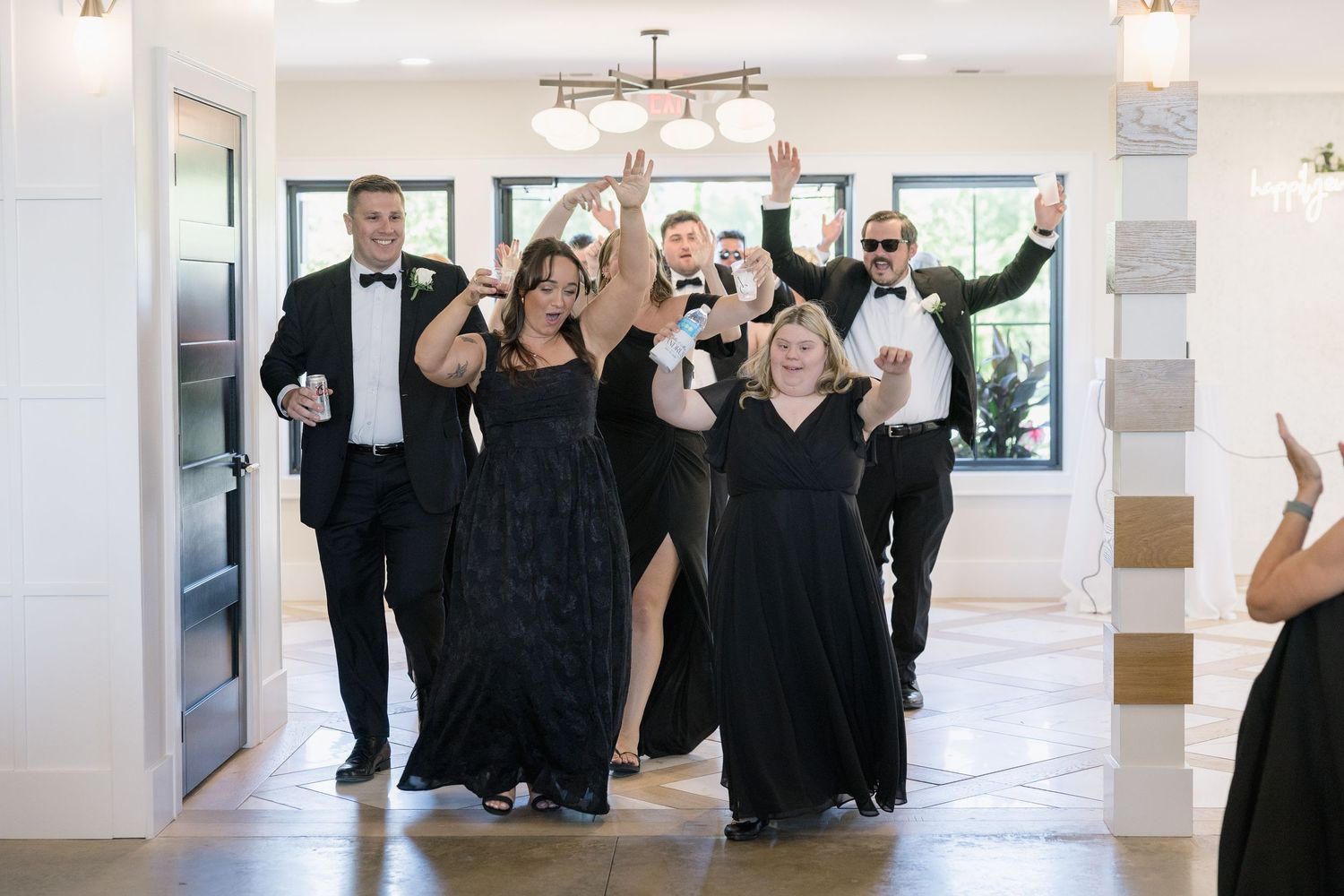 Wedding party in black formal attire makes grand entrance, celebrating with raised glasses and joyful expressions.