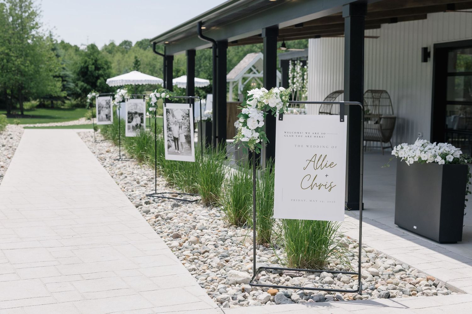 Outdoor wedding venue entrance with photo display panels and white floral arrangements lining pathway.