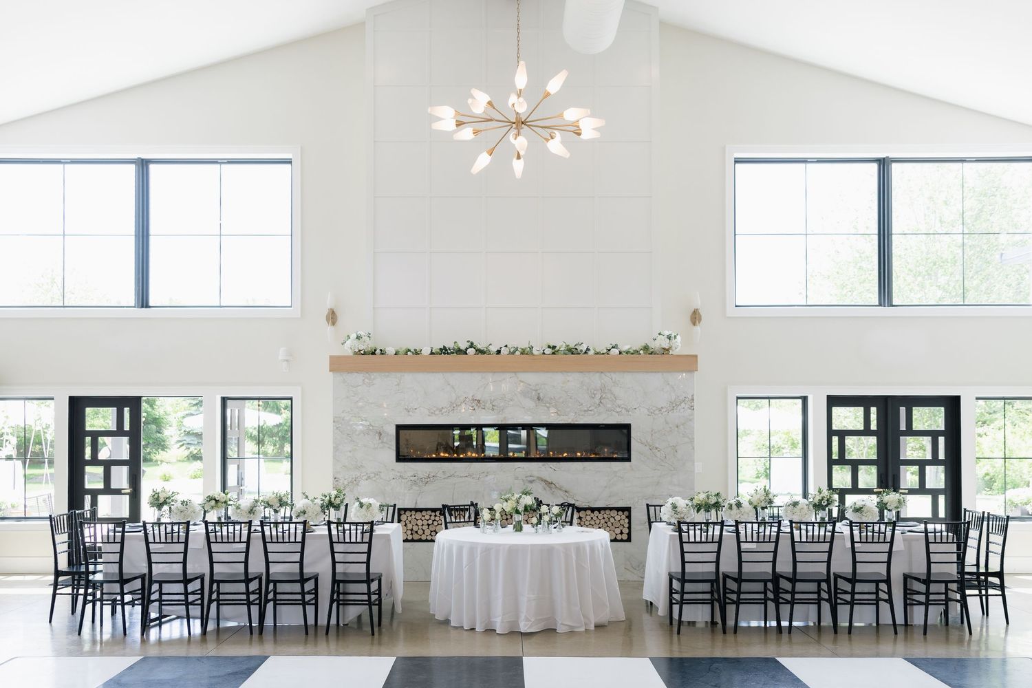 Elegant wedding reception hall with white vaulted ceilings, modern chandelier, fireplace, and black chiavari chairs at decorated tables.