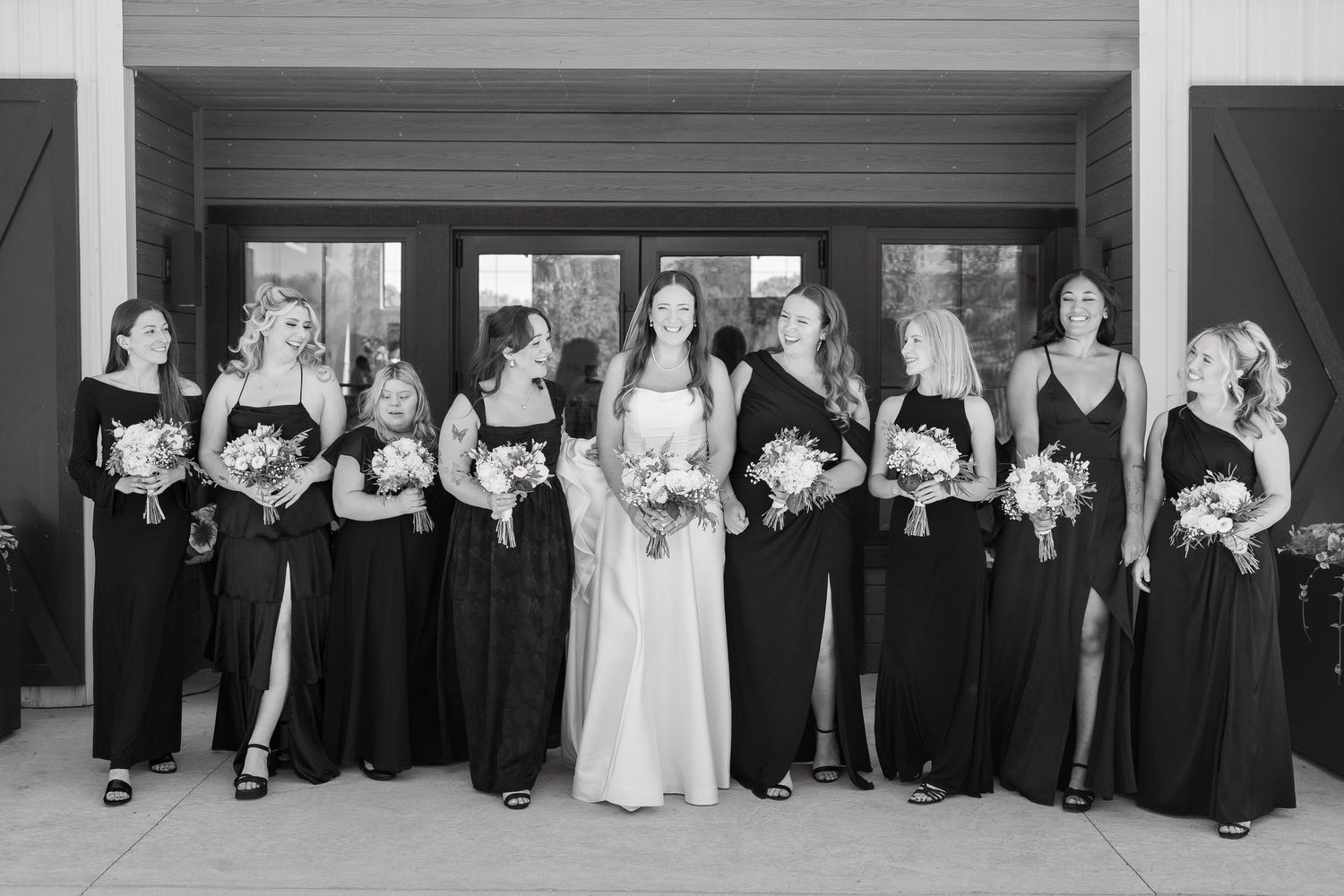 Black and white photo of a bride in white gown surrounded by bridesmaids in black dresses holding bouquets.