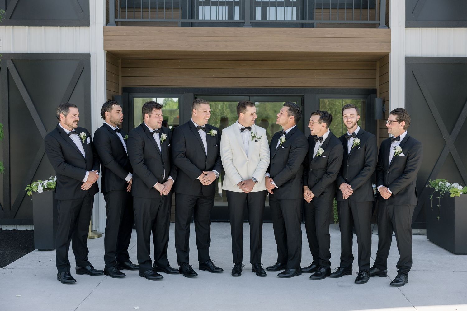 Groom in white tuxedo jacket stands with groomsmen in black tuxedos outside elegant wedding venue.