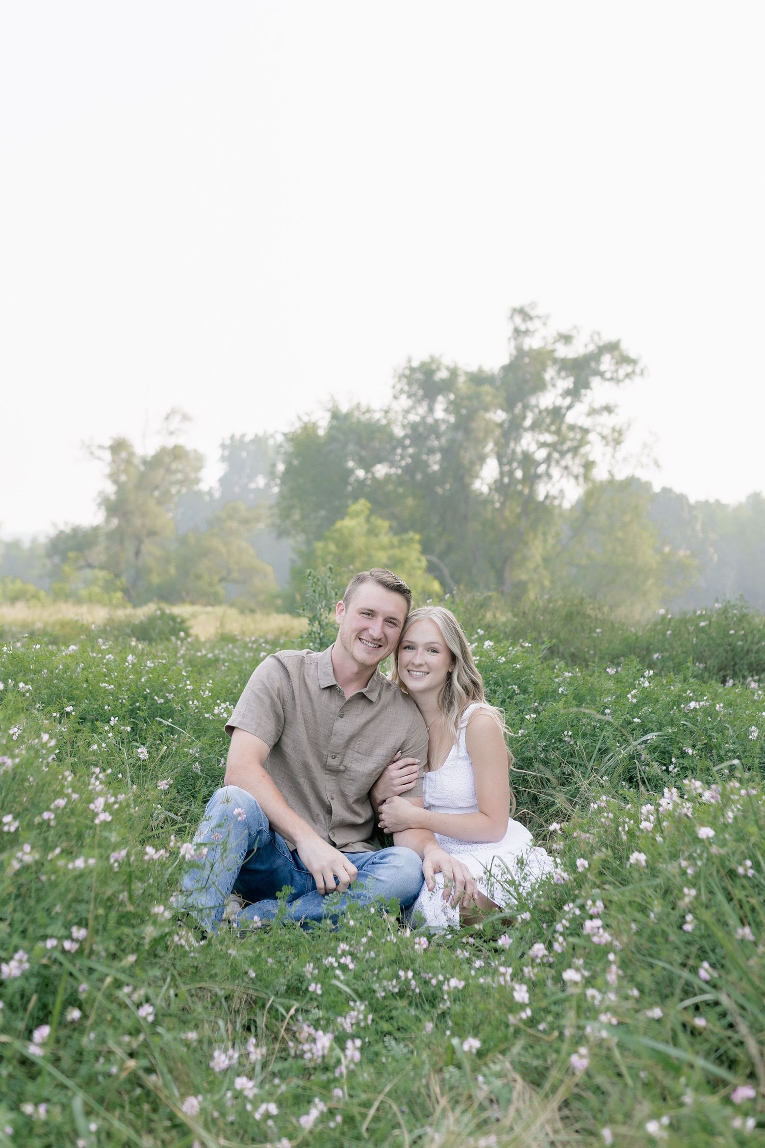 A couple sits together in a sunny wildflower meadow during a romantic outdoor photo session.