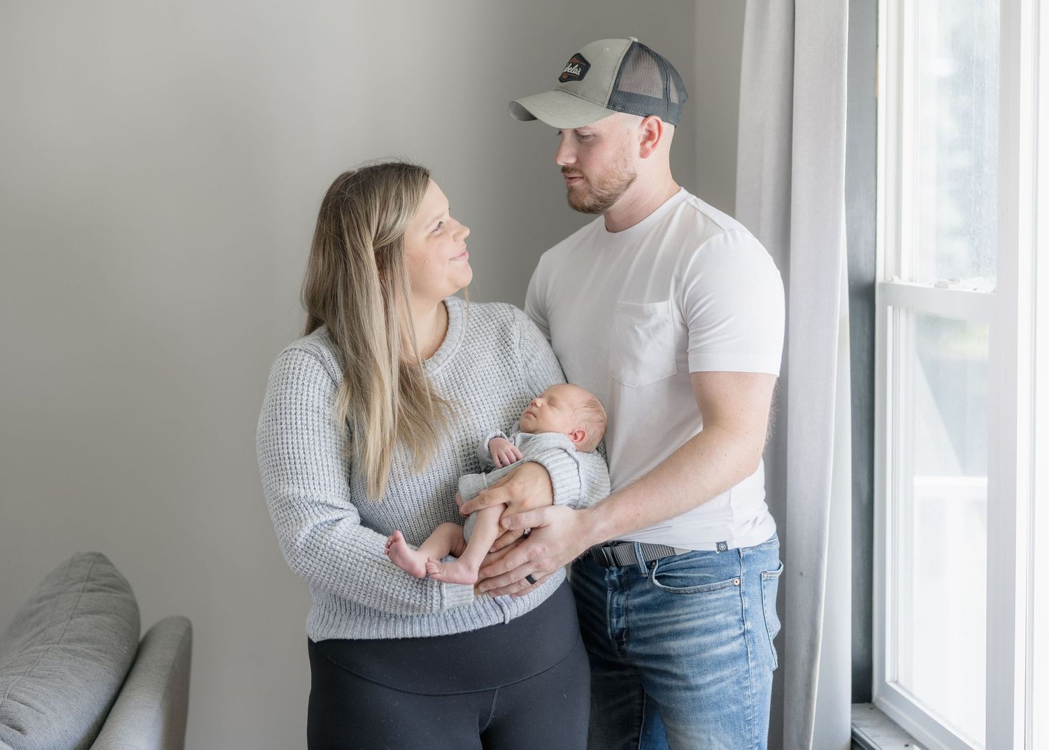 A couple stands lovingly near bright windows with their newborn baby in a modern minimalist home setting.