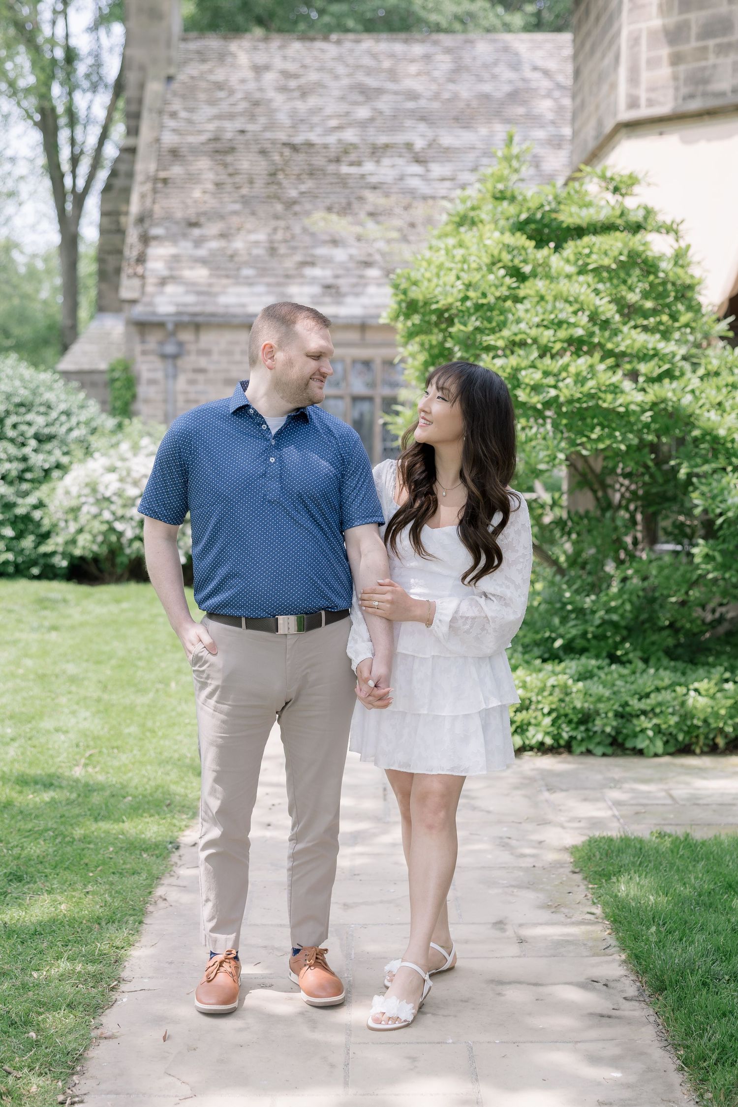 Couple poses on garden path in coordinated blue and white outfits near stone walls and greenery.