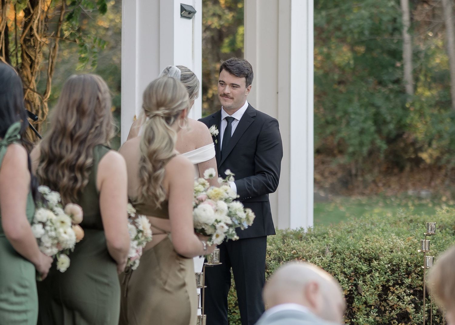 Wedding party gathers under an elegant white pergola with bridesmaids holding white bouquets.
