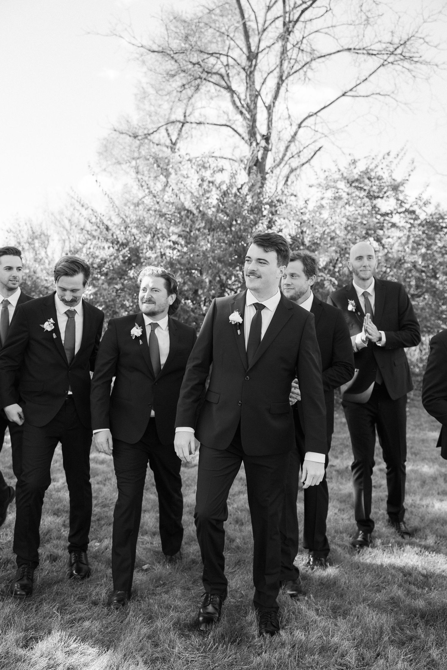 A group of groomsmen in black suits walk together on grass with bare trees in the background.
