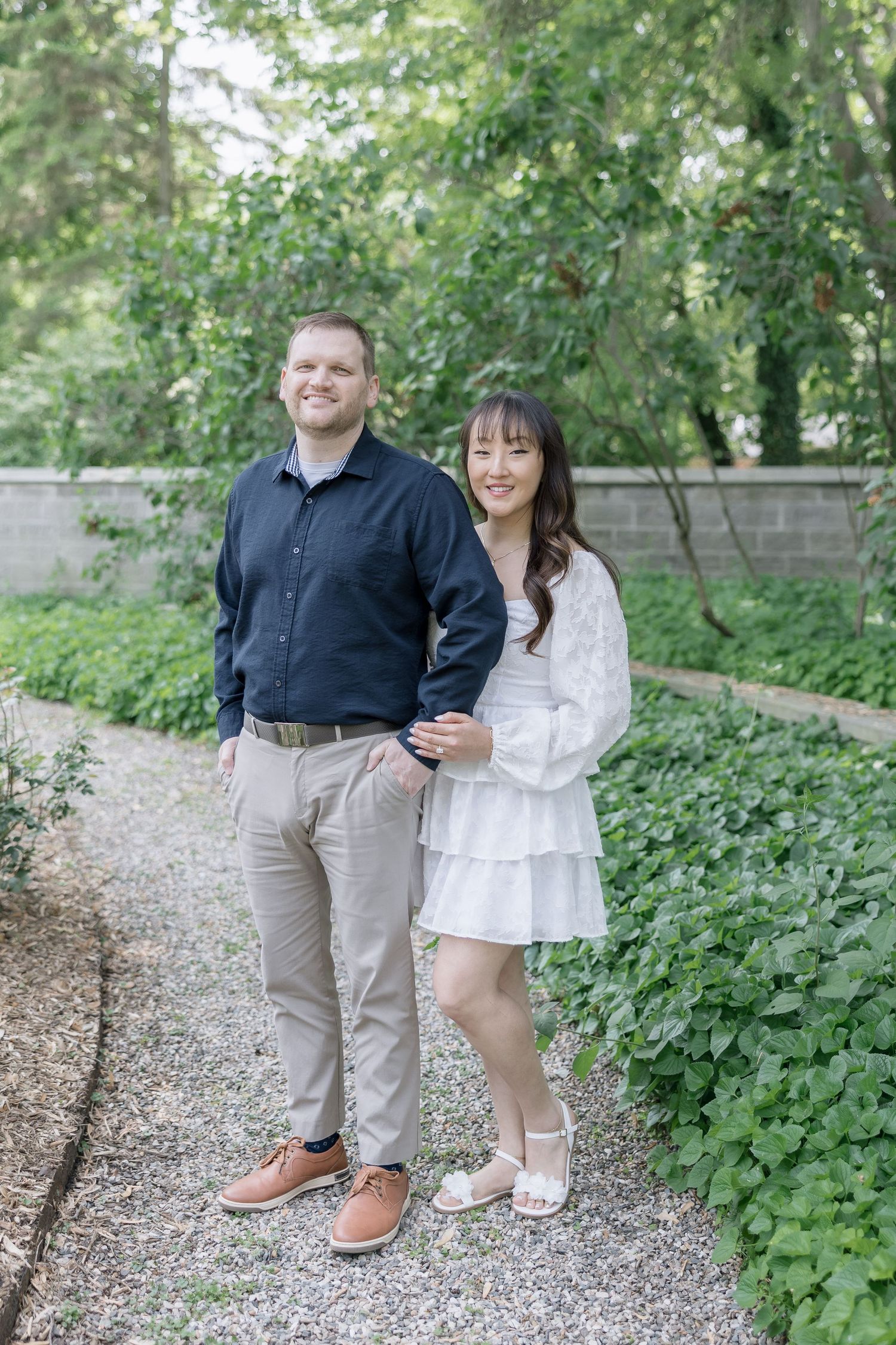 A couple poses together on a gravel path surrounded by lush green foliage in a garden setting.