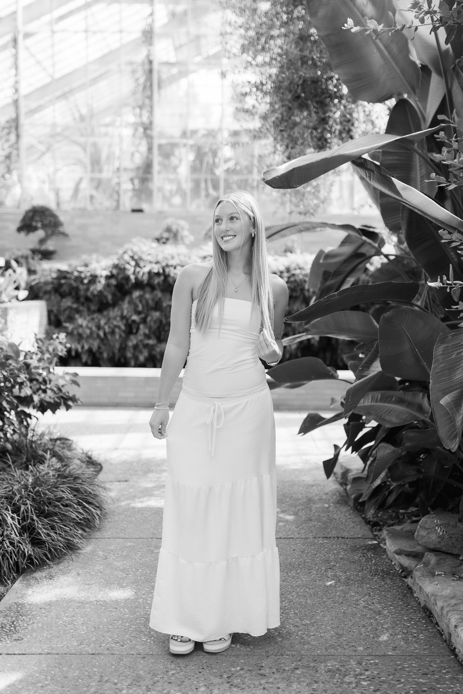 A young child in a white dress sits on a garden path while surrounded by lush foliage and plants.
