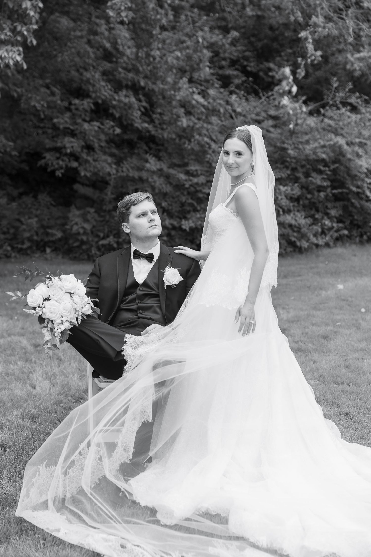 Black and white wedding photo of bride in veil and groom in tuxedo posing outdoors with bouquet.