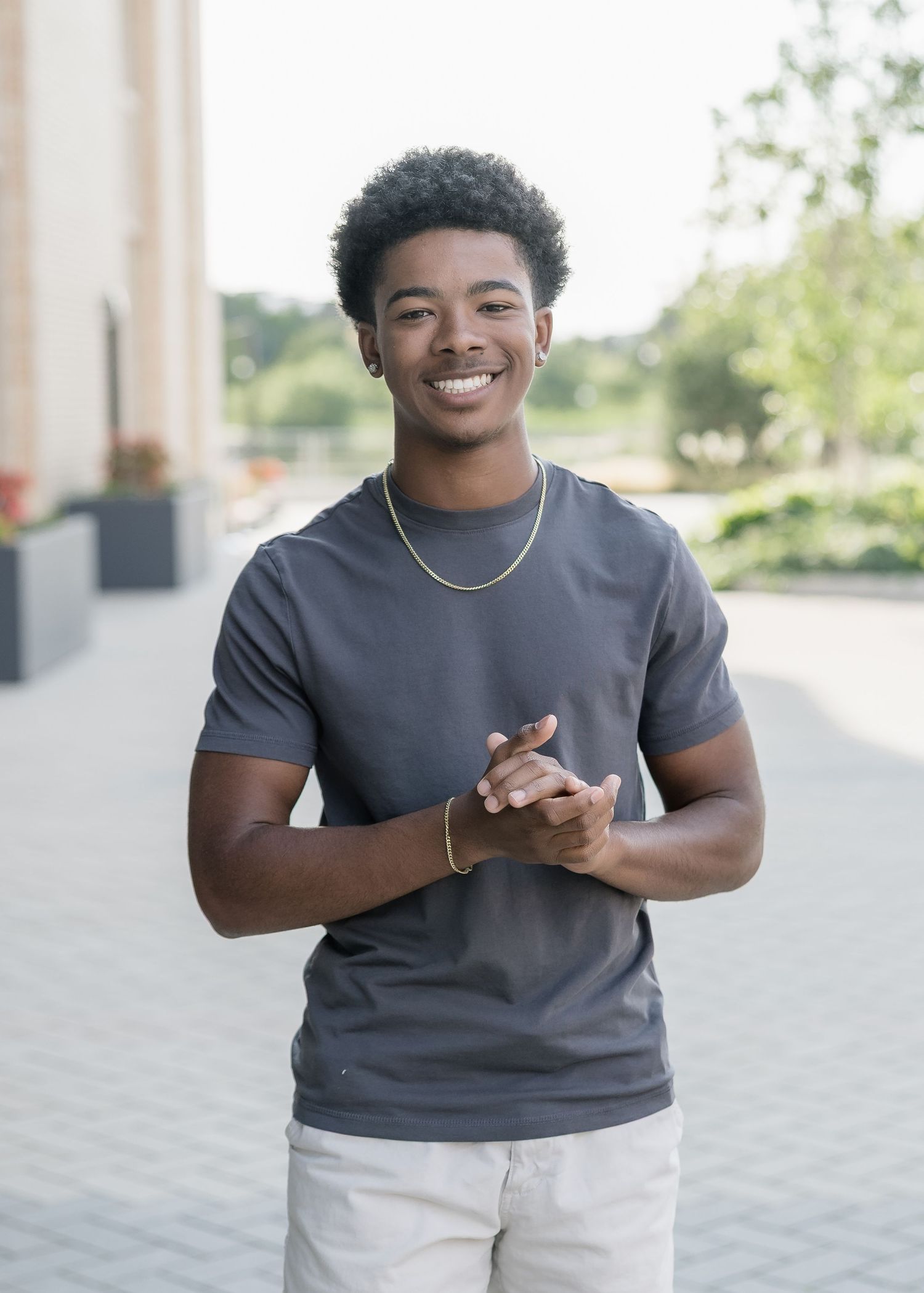 Young person in a gray t-shirt and white pants smiling and gesturing outdoors on a sunny day.