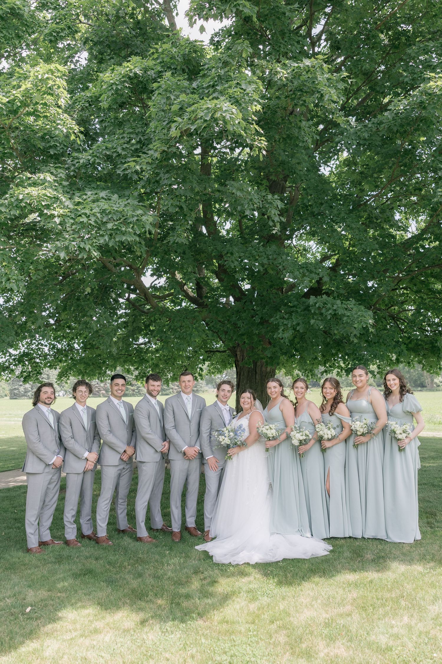 Wedding party in light gray attire poses together on grassy lawn under green summer trees.