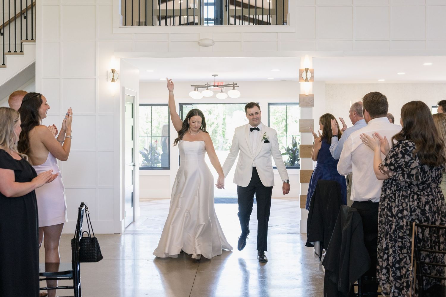 Bride and groom make grand entrance at wedding reception as guests applaud in elegant white venue.