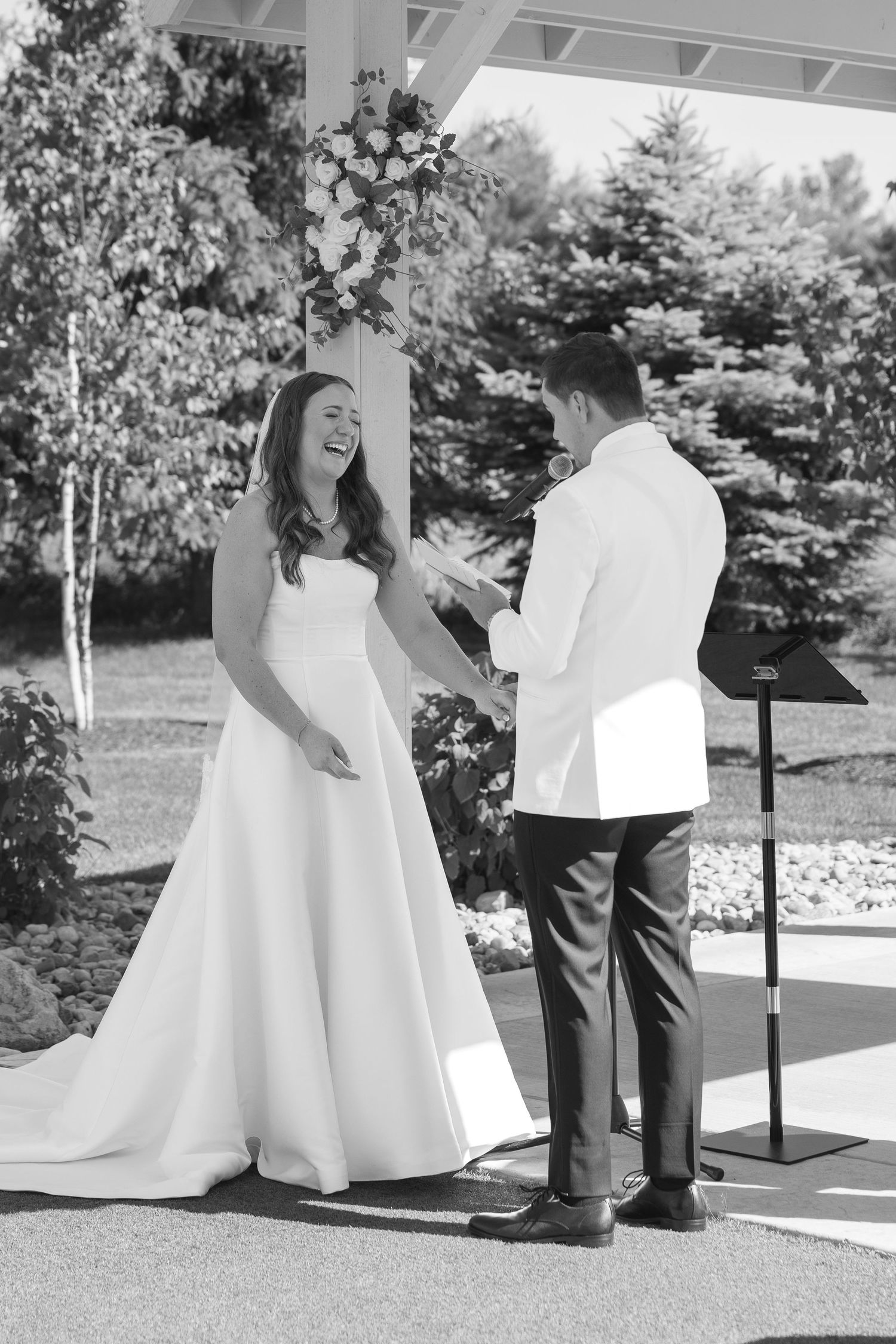 Black and white photo of bride laughing joyfully as groom reads vows at outdoor wedding ceremony.