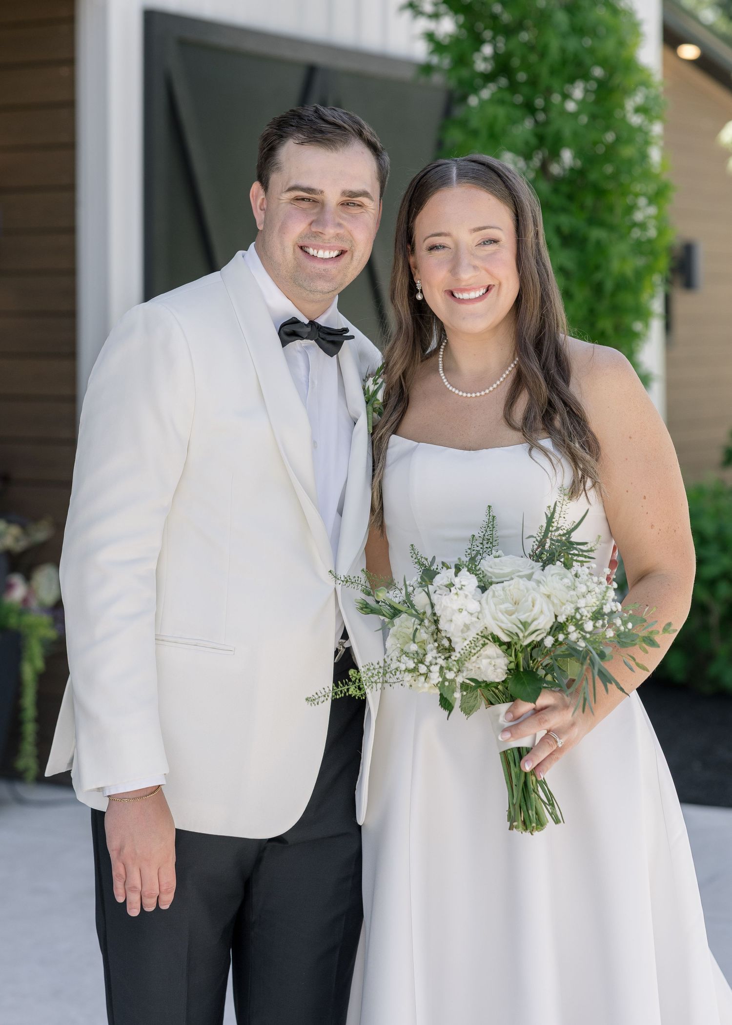 Smiling bride and groom pose together outdoors, she holds white floral bouquet, he wears white tuxedo jacket with bow tie.