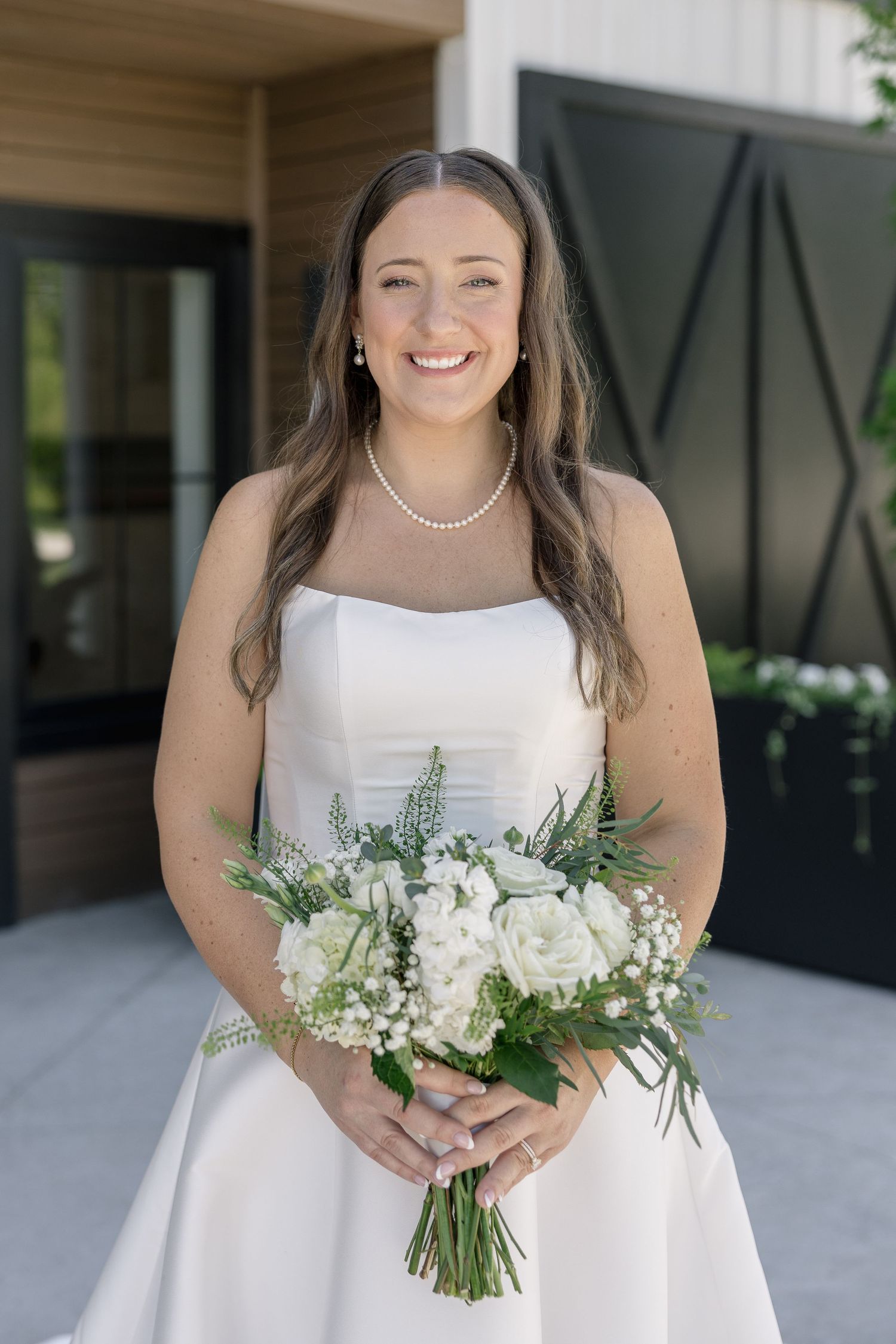 Smiling bride in a strapless white gown holds a white floral bouquet with greenery, wearing a pearl necklace.