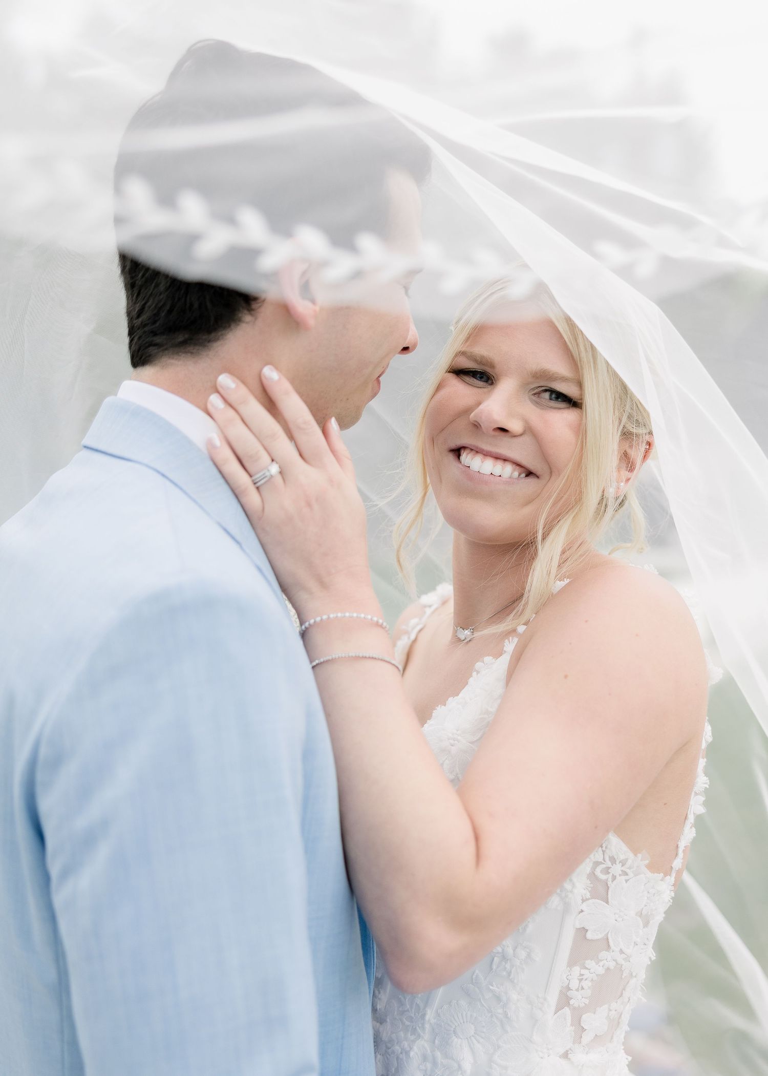 A smiling bride in a lace gown gazes at her groom under a flowing veil during an outdoor wedding ceremony.