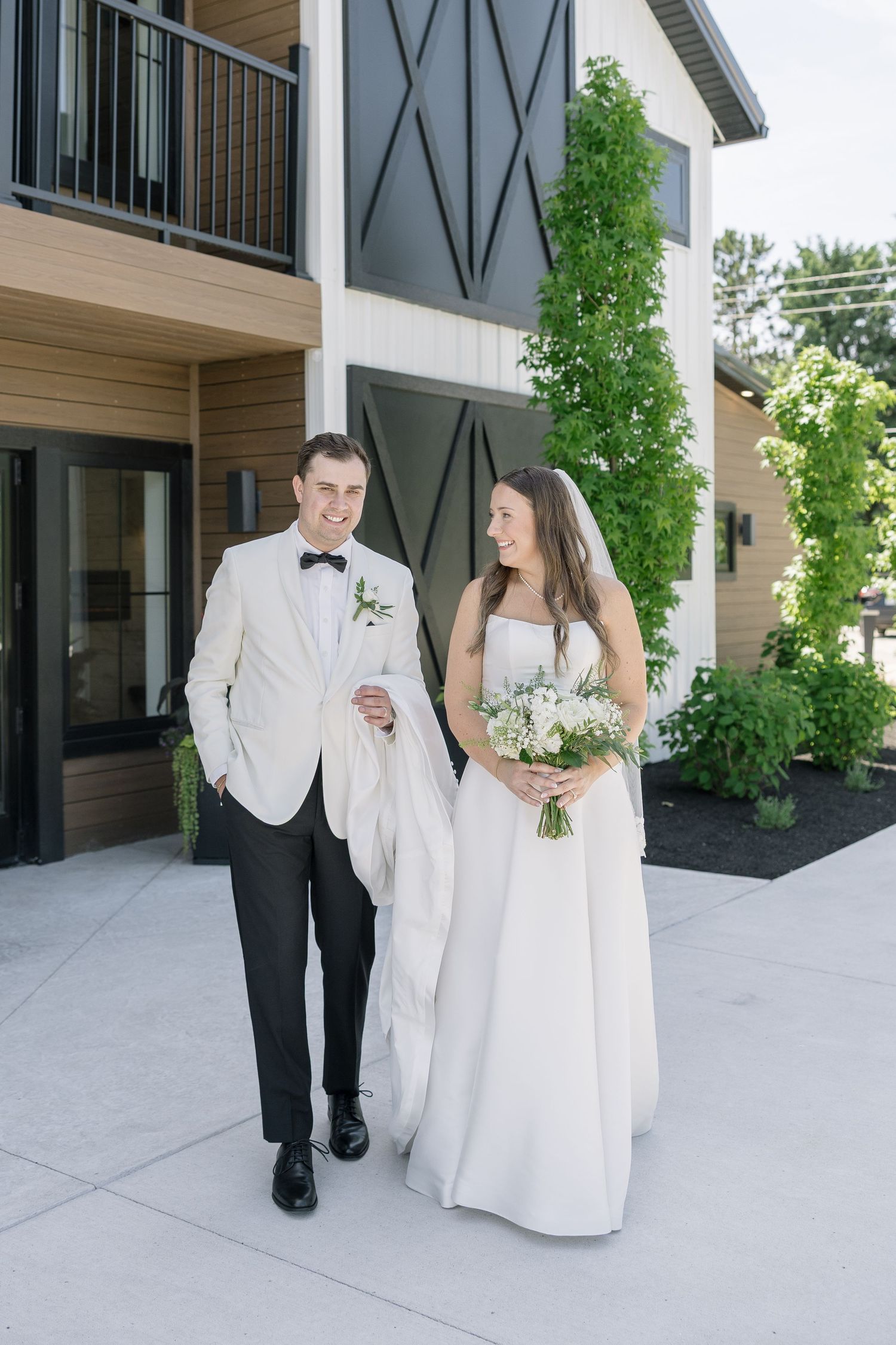 Bride and groom walking together outside a modern barn venue, bride holding white bouquet, groom in ivory tuxedo jacket.