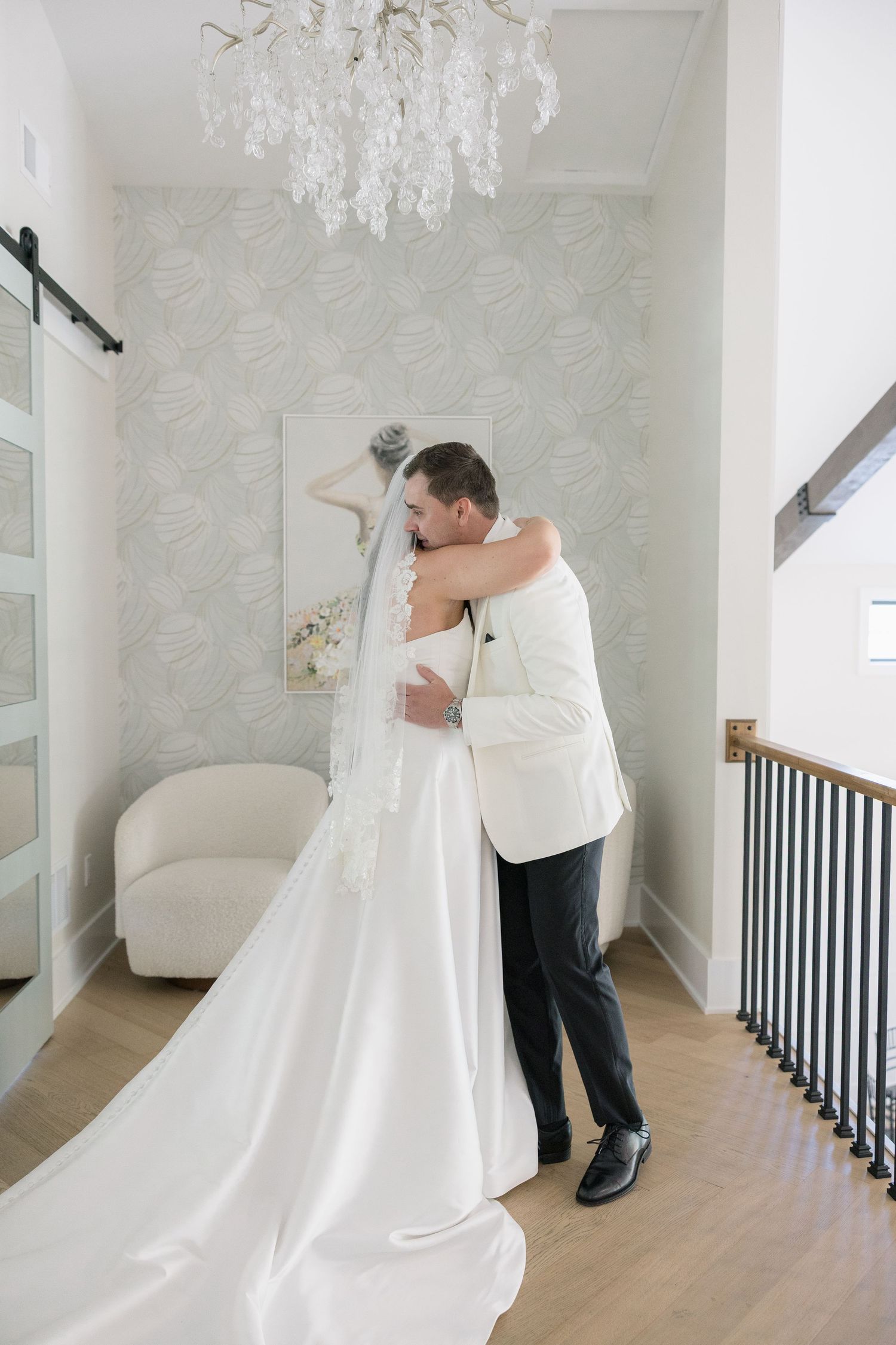 Bride and groom share a tender embrace in an elegant white interior with chandelier on their wedding day.