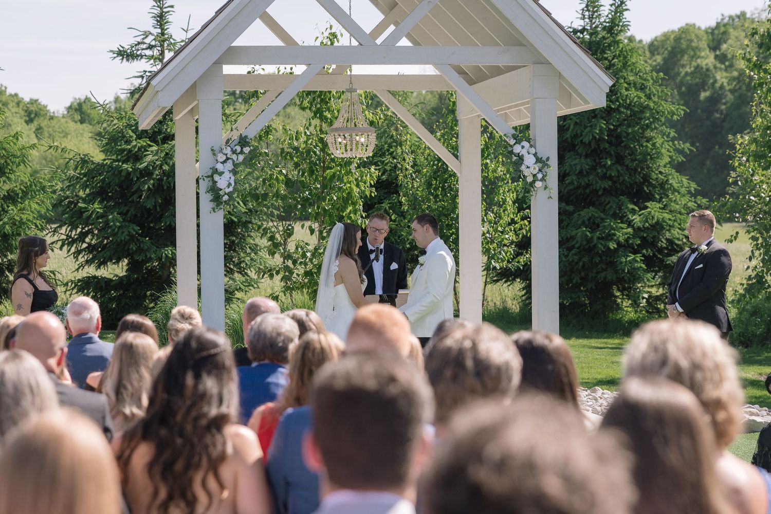 Outdoor wedding ceremony under white gazebo with chandelier, couple exchanging vows before guests on lush green grounds.