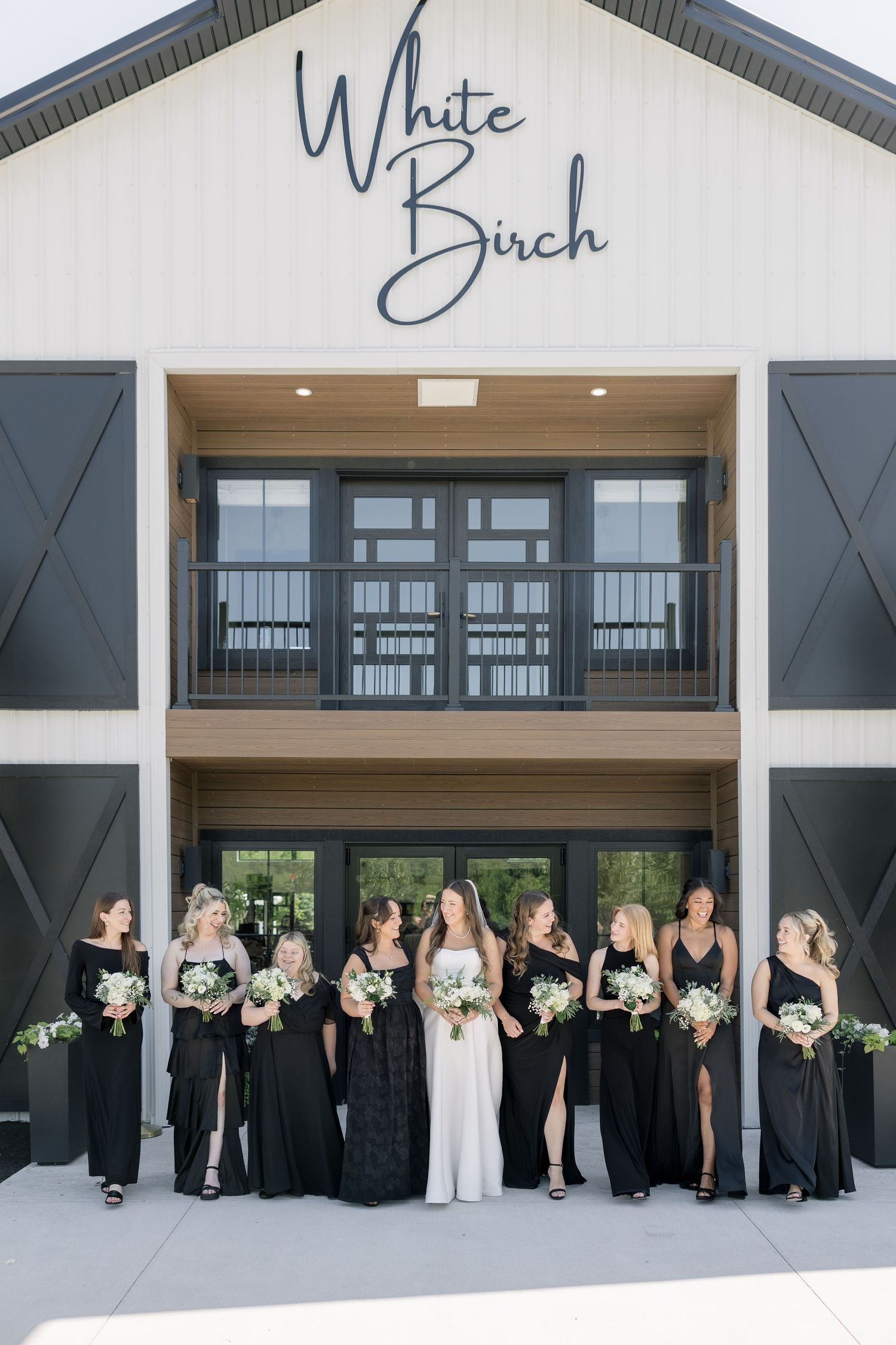Bride in white gown with bridesmaids in black dresses holding white bouquets outside White Birch wedding venue.