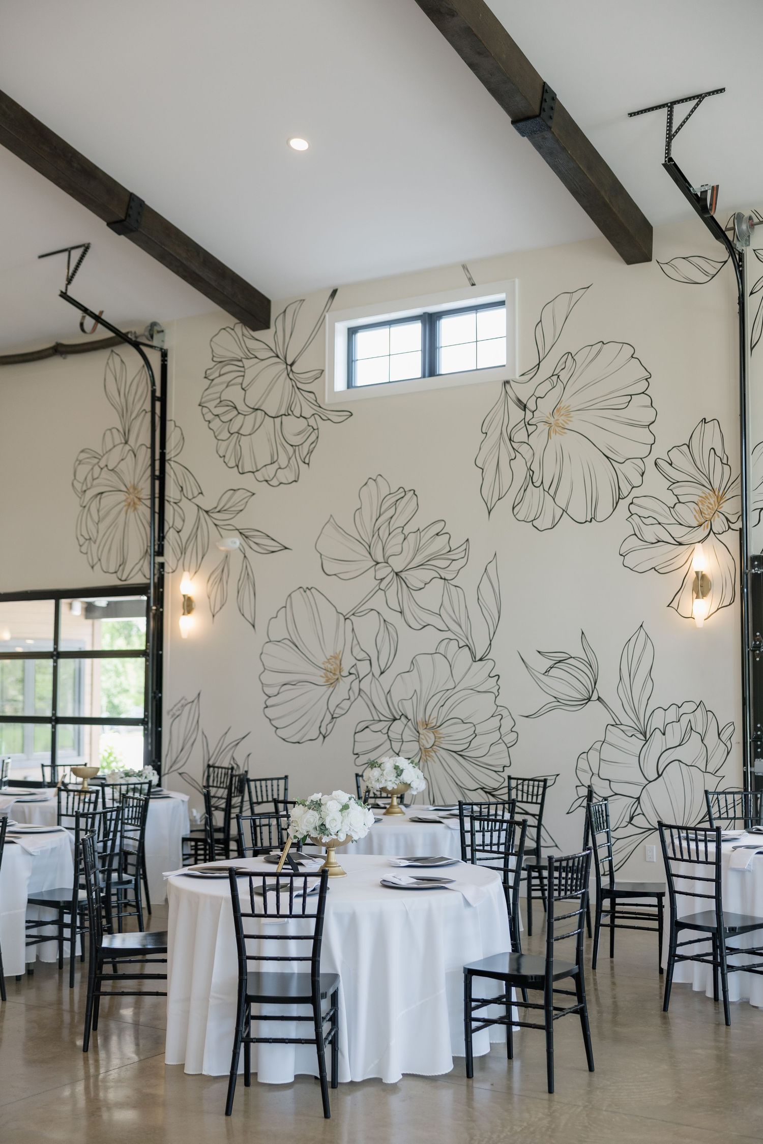 Bright wedding reception hall with large floral wall mural, black chiavari chairs, and white round tables with centerpieces.