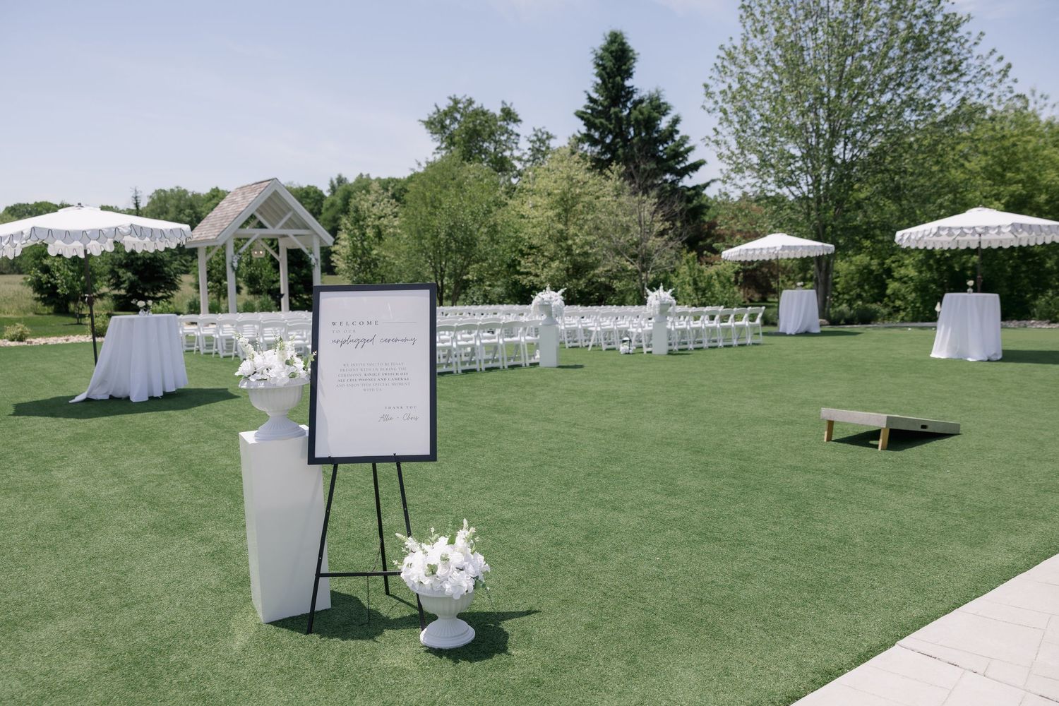 Elegant outdoor wedding ceremony setup with white chairs, floral arrangements, gazebo, and welcome sign on lush green lawn.
