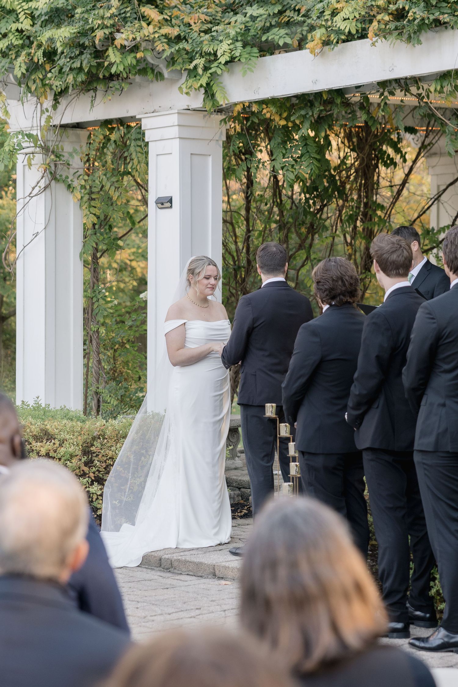 An outdoor wedding ceremony taking place under a white pergola with climbing greenery.