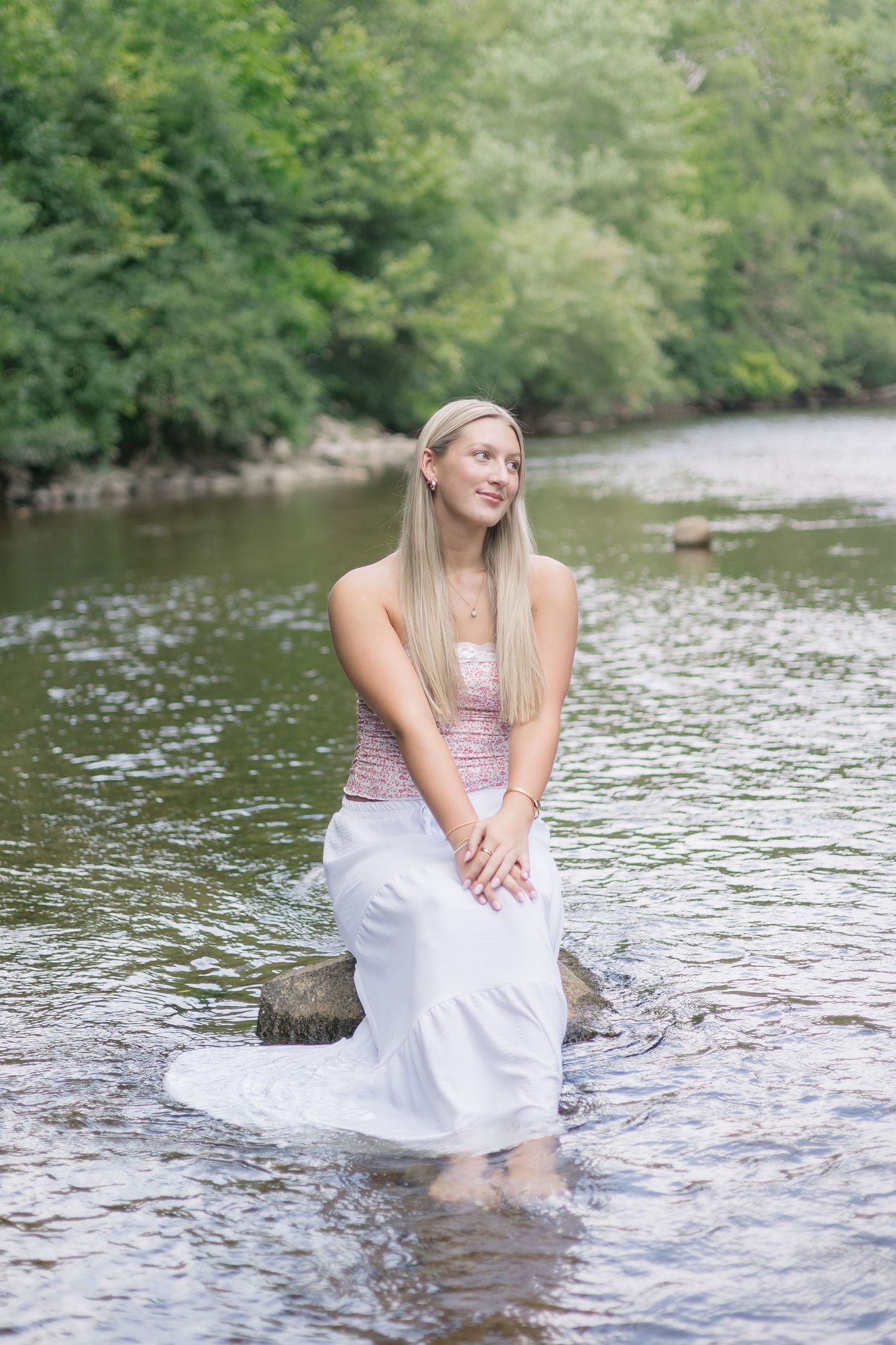 A person in a white dress sits gracefully in a flowing stream surrounded by lush green trees and rocks.
