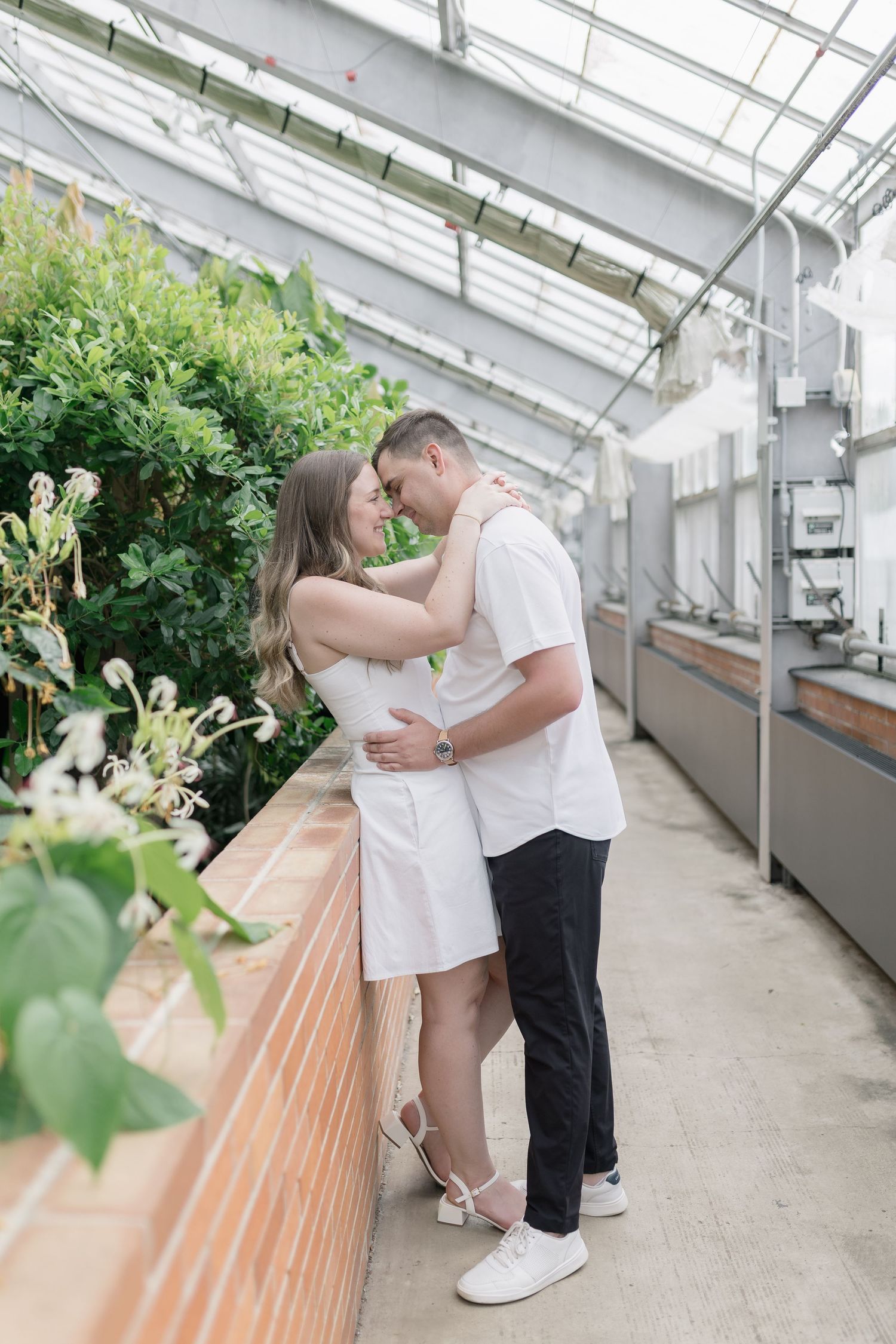 Romantic couple shares tender moments in a bright greenhouse with white floors and lush greenery.